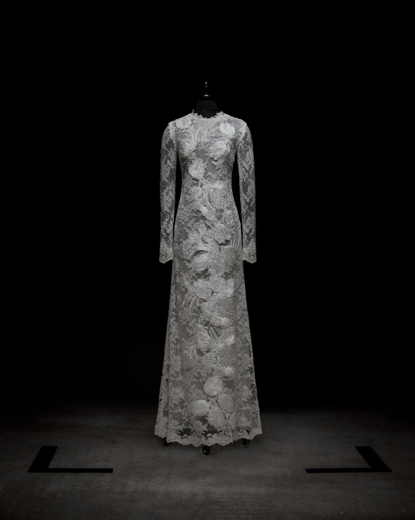 El vestido Dava es un vestido de encaje blanco de manga larga con estampado floral y transparencias. De talle alto y escote pronunciado, se exhibe hasta el suelo sobre un maniquí negro sobre un fondo oscuro.