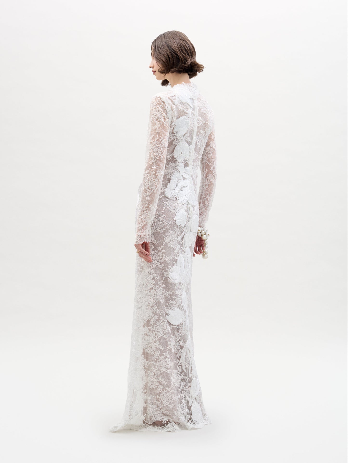 Una persona con el vestido Dava —un traje largo y transparente de encaje blanco con estampados florales— se encuentra de pie sobre un fondo blanco liso, ligeramente girada hacia un lado. Tiene el pelo castaño y corto.