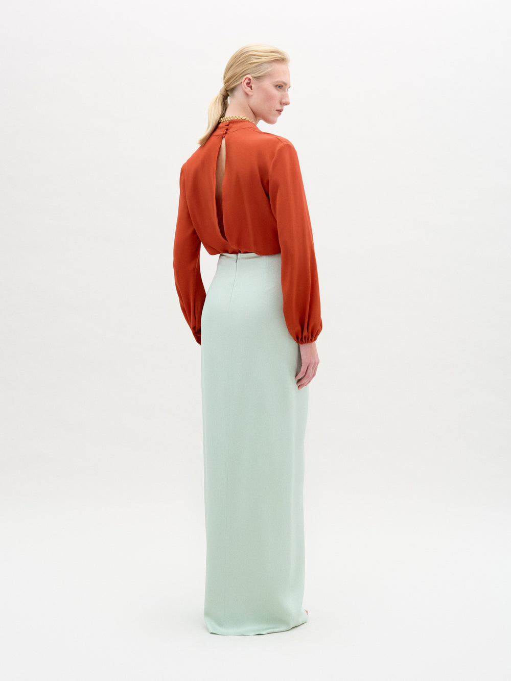 Una mujer mira hacia otro lado, con una blusa de manga larga de color óxido con abertura en la espalda y la falda Gadea -una maxi de talle alto de color verde menta claro confeccionada en tejido italiano- sobre un fondo claro liso.