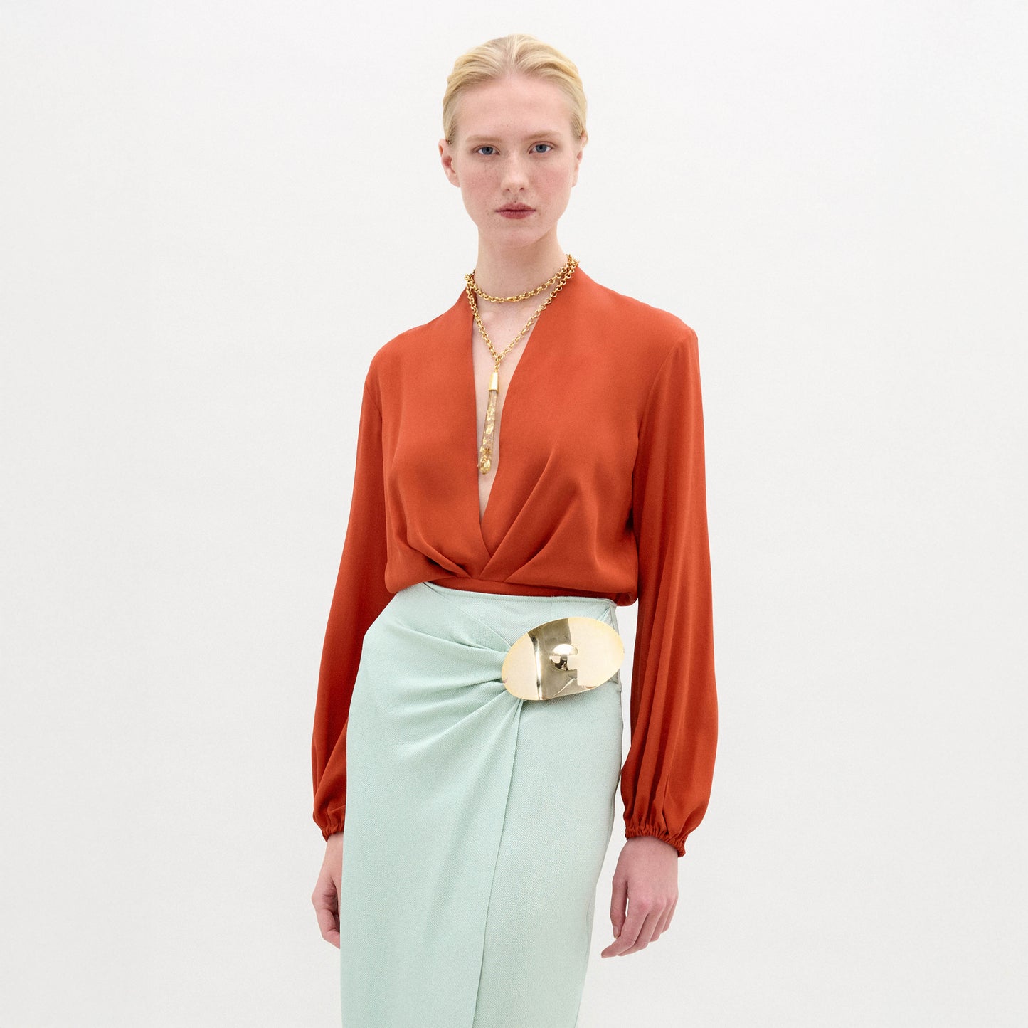 Una persona con el pelo rubio recogido en un moño bajo lleva la blusa Delfina de color óxido, confeccionada en seda 100%, en forma de top envolvente de manga larga, combinada con una falda de color menta pálido y un cinturón metálico dorado sobre un fondo blanco liso.