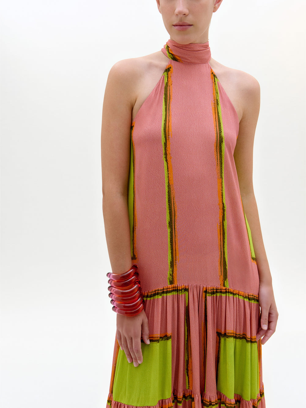 Una persona lleva el vestido Dorelia, un maxi vestido sin mangas con escote halter, paneles rosas y verdes y rayas verticales naranjas y amarillas. Sobre un fondo liso, lleva un brazalete rosa en la muñeca izquierda. Rostro parcialmente fuera de cuadro.