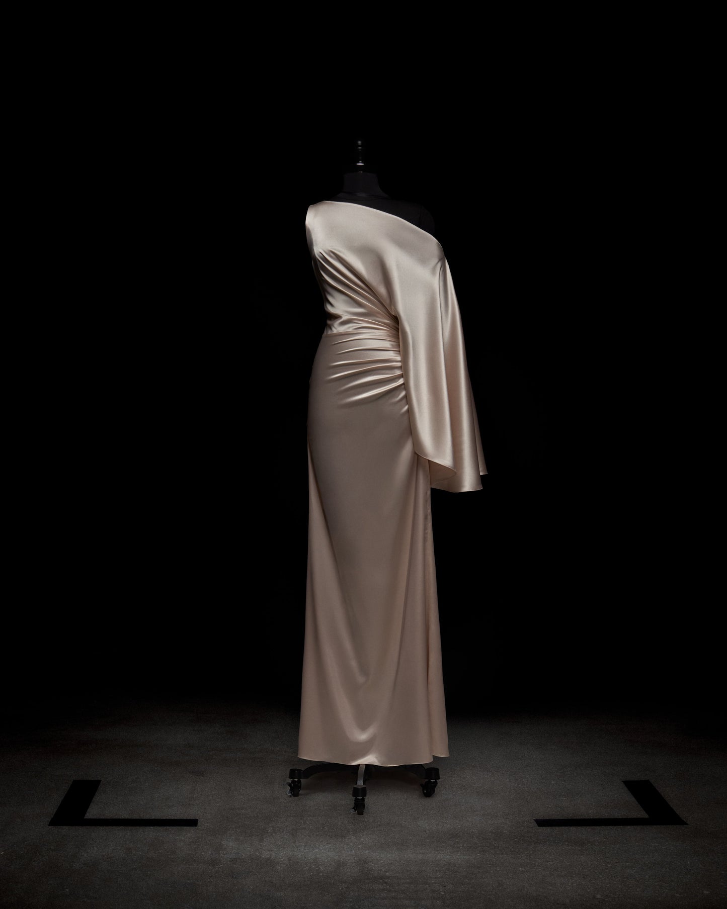 El Vestido Dotty, un vestido de noche largo de satén color champán con un diseño asimétrico y una manga, se exhibe sobre un maniquí negro sobre un fondo oscuro.