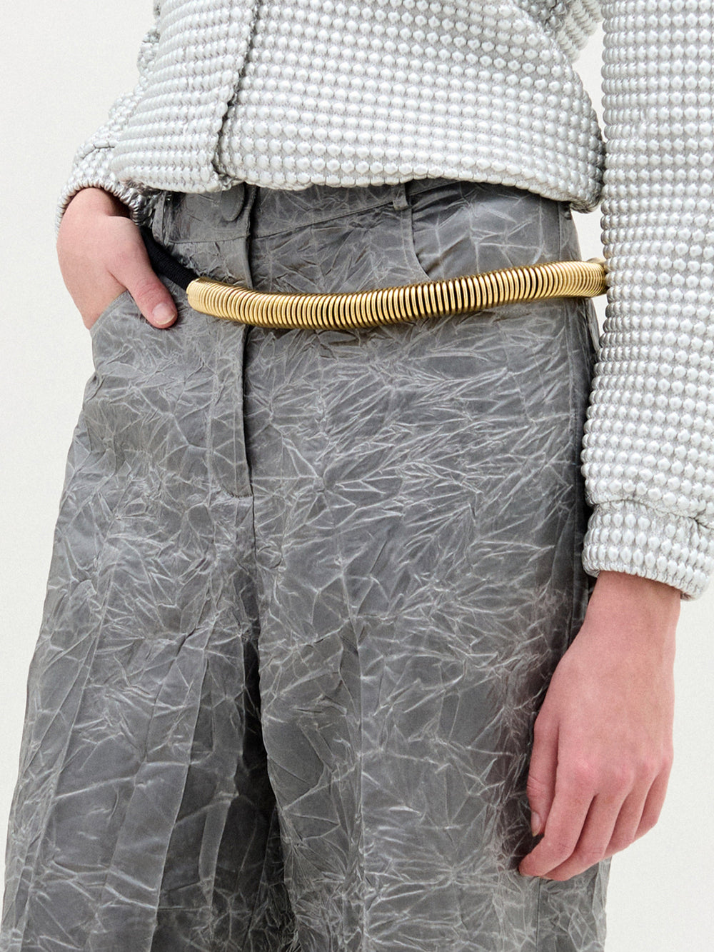 Con unos pantalones grises arrugados y una camisa blanca texturizada con pequeños lunares en relieve, esta persona lleva como accesorio el cinturón Dunia, un grueso cinturón dorado hecho a mano en Colombia, que destaca mientras su mano izquierda descansa en un bolsillo.