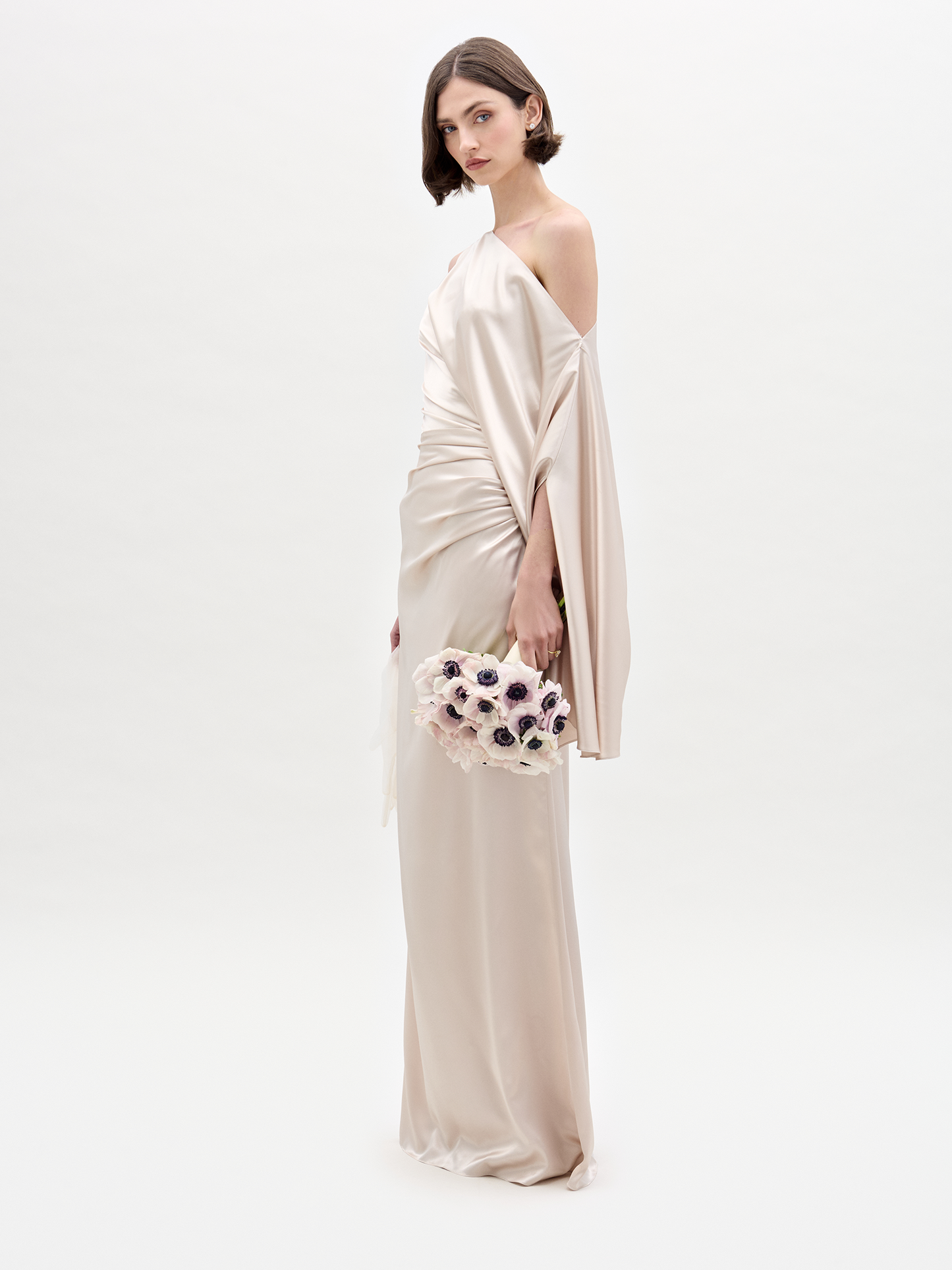 Una mujer que viste el Vestido Dotty, un vestido largo de seda color champán con hombros descubiertos, sostiene un ramo de flores blancas y violetas sobre un fondo blanco liso.
