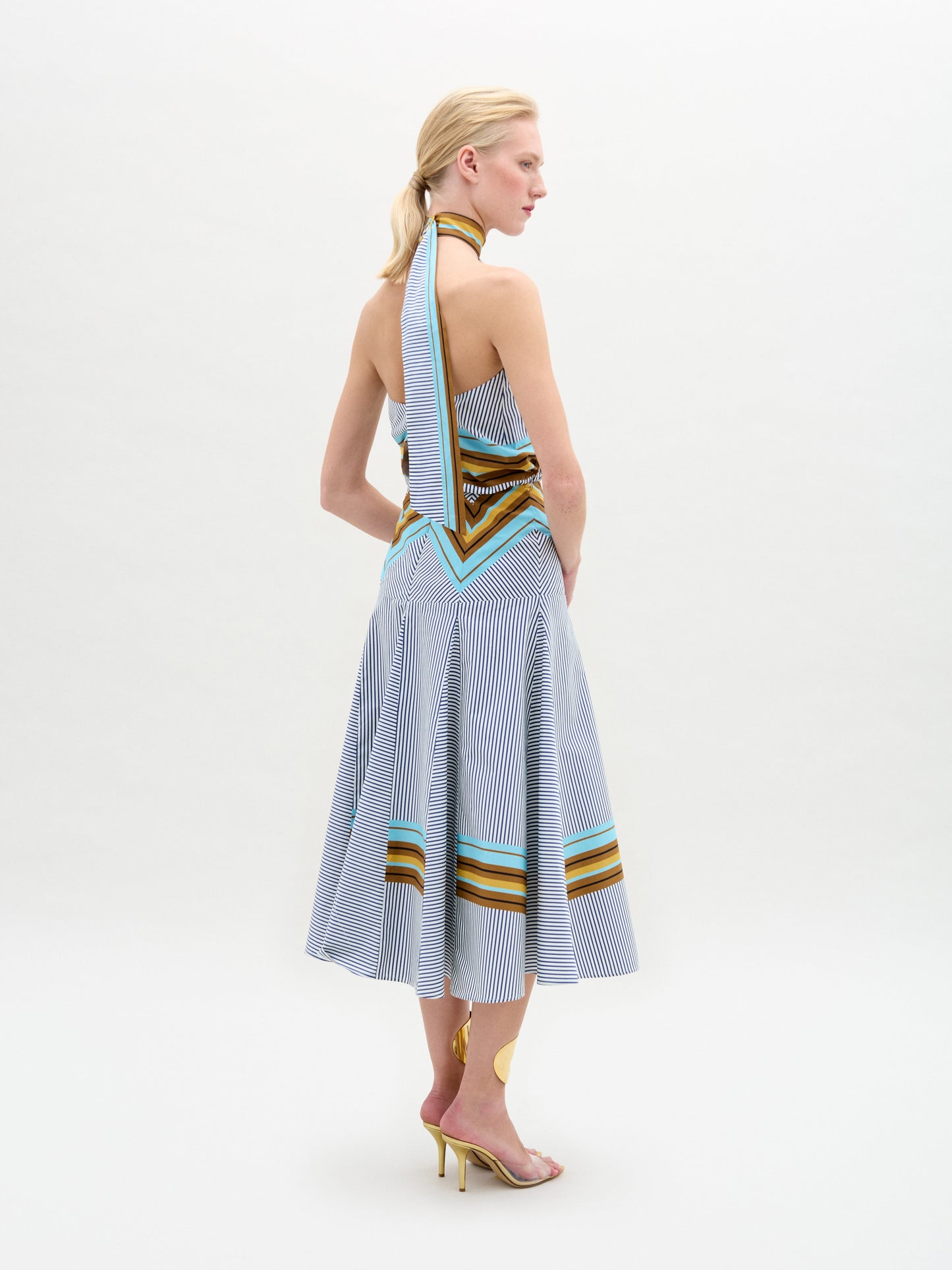 Una mujer rubia con coleta baja lleva el vestido Elara, un midi a rayas azules y blancas hecho en Colombia con detalles en marrón, amarillo y turquesa, combinado con tacones dorados. Está de pie ante un fondo blanco liso.