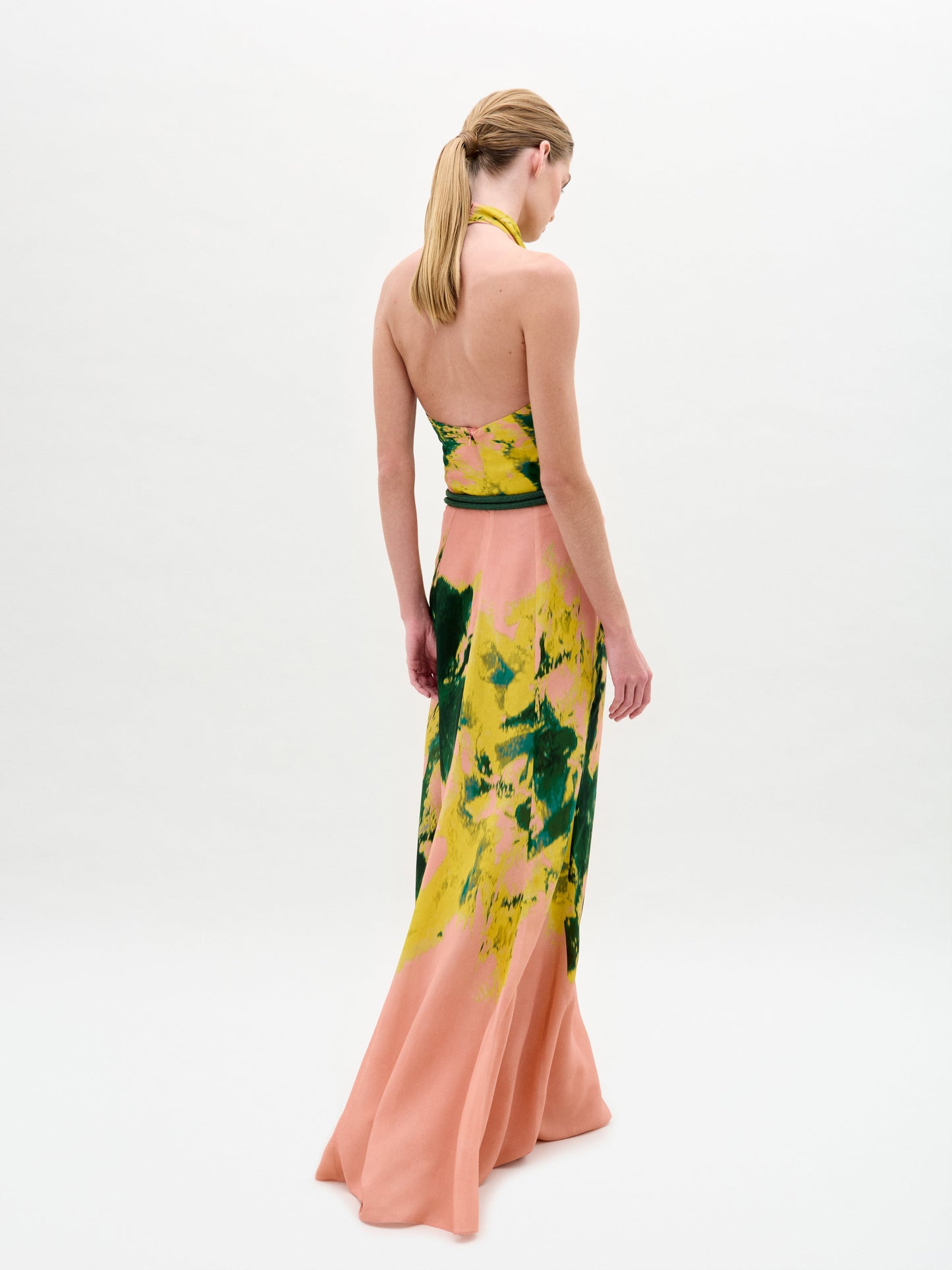 Una mujer con el pelo rubio recogido en una coleta lleva el Ellis Dress -un maxi de tela italiana con base melocotón y estampado abstracto amarillo-verde- de espaldas a la cámara sobre un fondo blanco liso.