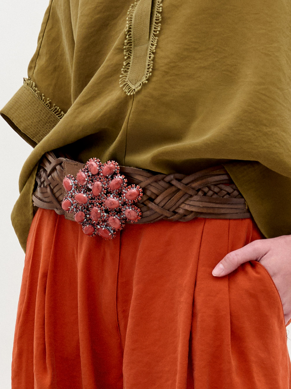 Una persona lleva un top verde oliva con textura y unos pantalones naranjas de cintura alta, adornados con el Cinturón Eines -un cinturón de cuero italiano trenzado marrón con una gran hebilla floral rosa ornamentada- y la mano derecha en el bolsillo.