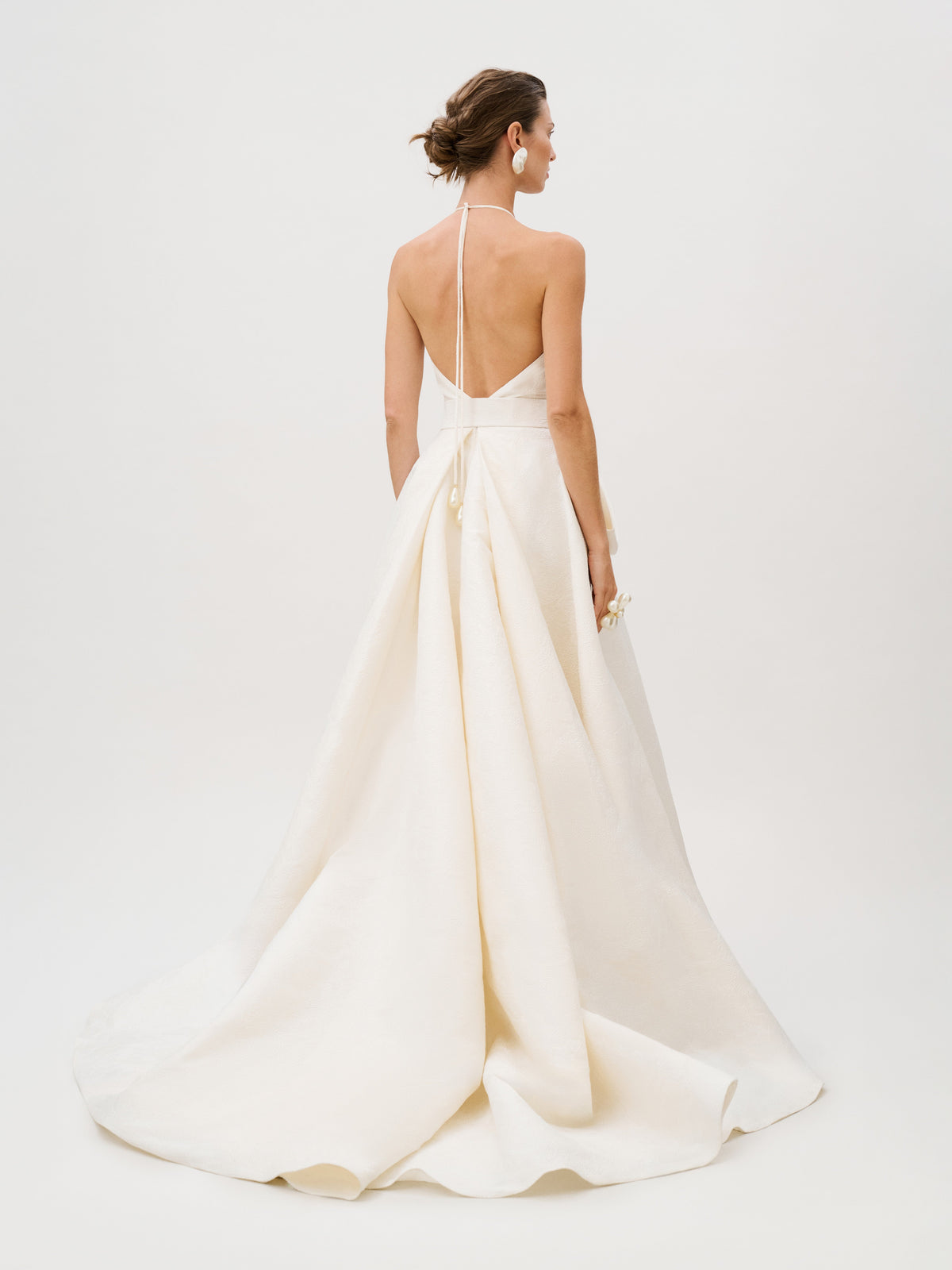 A woman stands facing away, holding a small bouquet, wearing the Elsa Dress—a white halter-neck, backless gown crafted from luxurious Italian fabric with a long train.