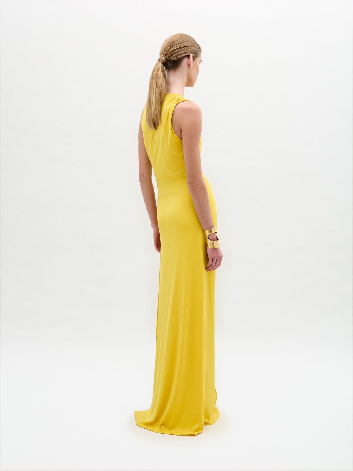 Una mujer con el pelo rubio recogido en una coleta baja está de espaldas, con el vestido amarillo Fadila con detalles dorados y brazaletes de oro, sobre un fondo blanco liso.