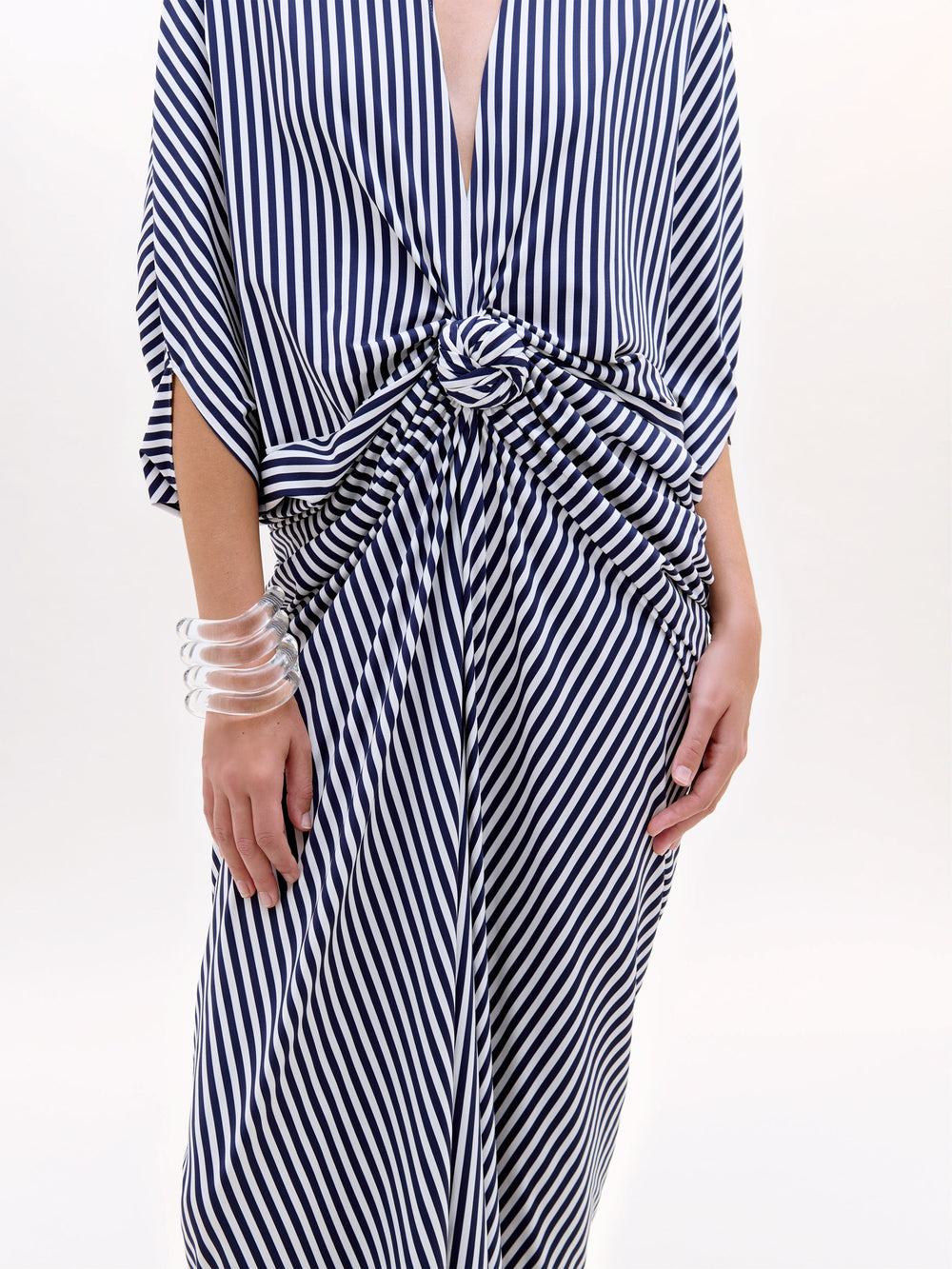 Se ve a una persona con la Túnica Fiore, un maxivestido de rayas verticales azules y blancas con cintura de nudo retorcido, escote en V y un brazalete de trozos transparentes. No se ve la cabeza; el fondo es liso y claro.