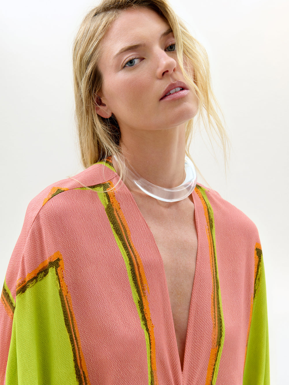 Una mujer de pelo largo y rubio lleva la Túnica Fiore, una maxi holgada en rosa y verde con toques naranjas, confeccionada en tejido italiano. La combina con un collar grueso transparente y mira hacia arriba sobre un fondo liso.