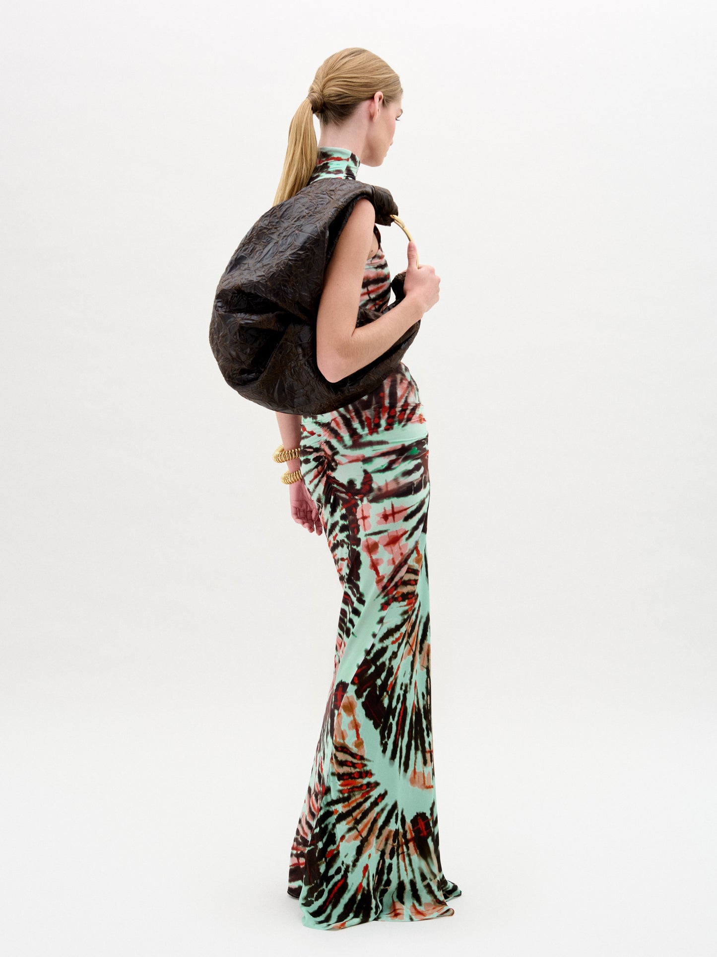 Una persona aparece de perfil sobre un fondo blanco liso, con el vestido Frances -un maxi ajustado sin mangas en rojo, negro y verde-, brazaletes dorados y un gran bolso negro sobre el hombro.