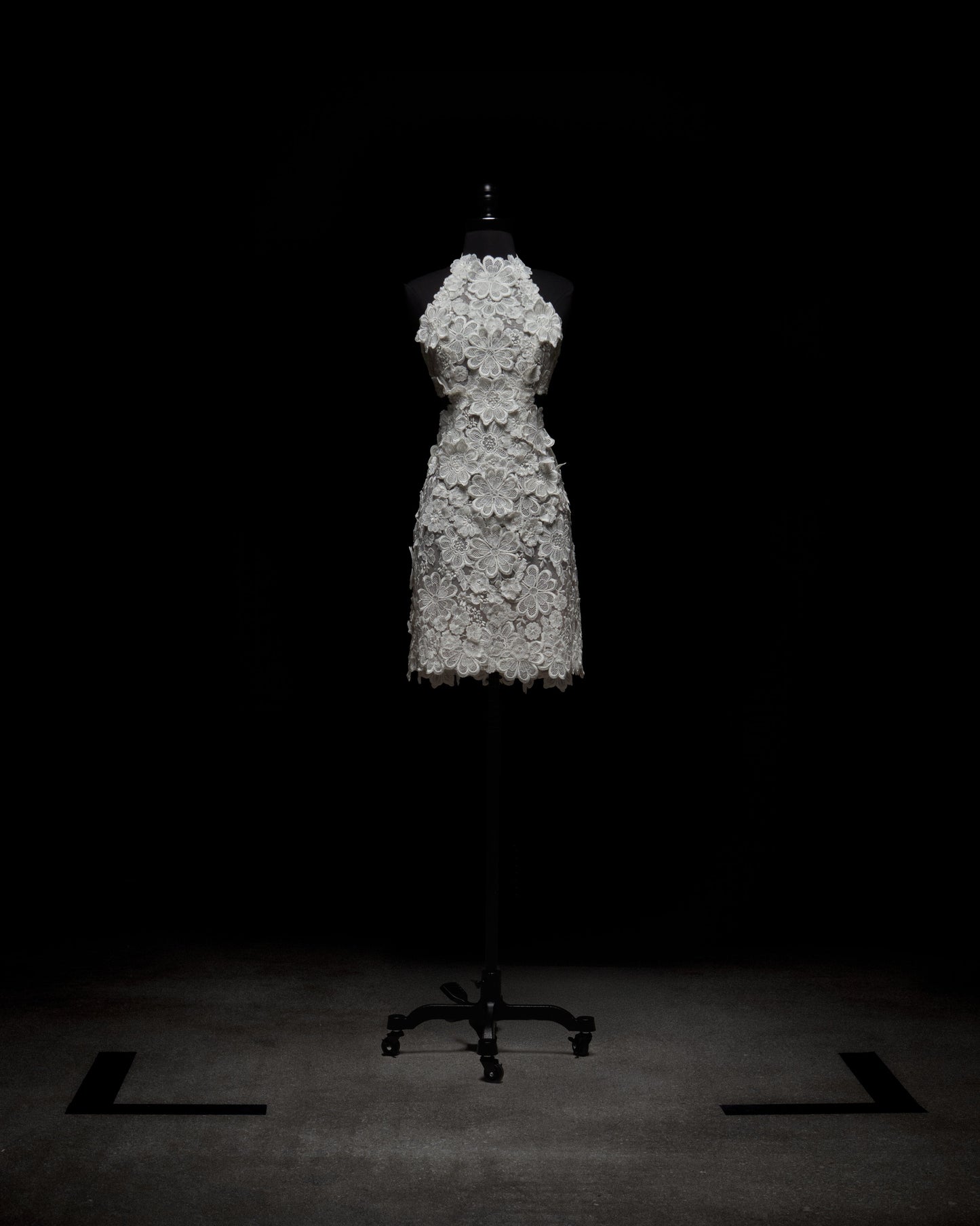 El vestido Garazi, un diseño de encaje blanco con escote halter y patrones florales intrincados, se exhibe en un maniquí negro sobre un fondo oscuro.