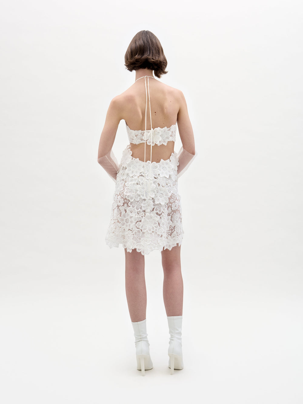 Una persona de cabello castaño corto está de espaldas a la cámara con un vestido Garazi, un diseño de encaje blanco sin mangas ni espalda descubierta con estampados florales. Completa el look con botas blancas de tacón y guantes transparentes sobre un fondo blanco liso.