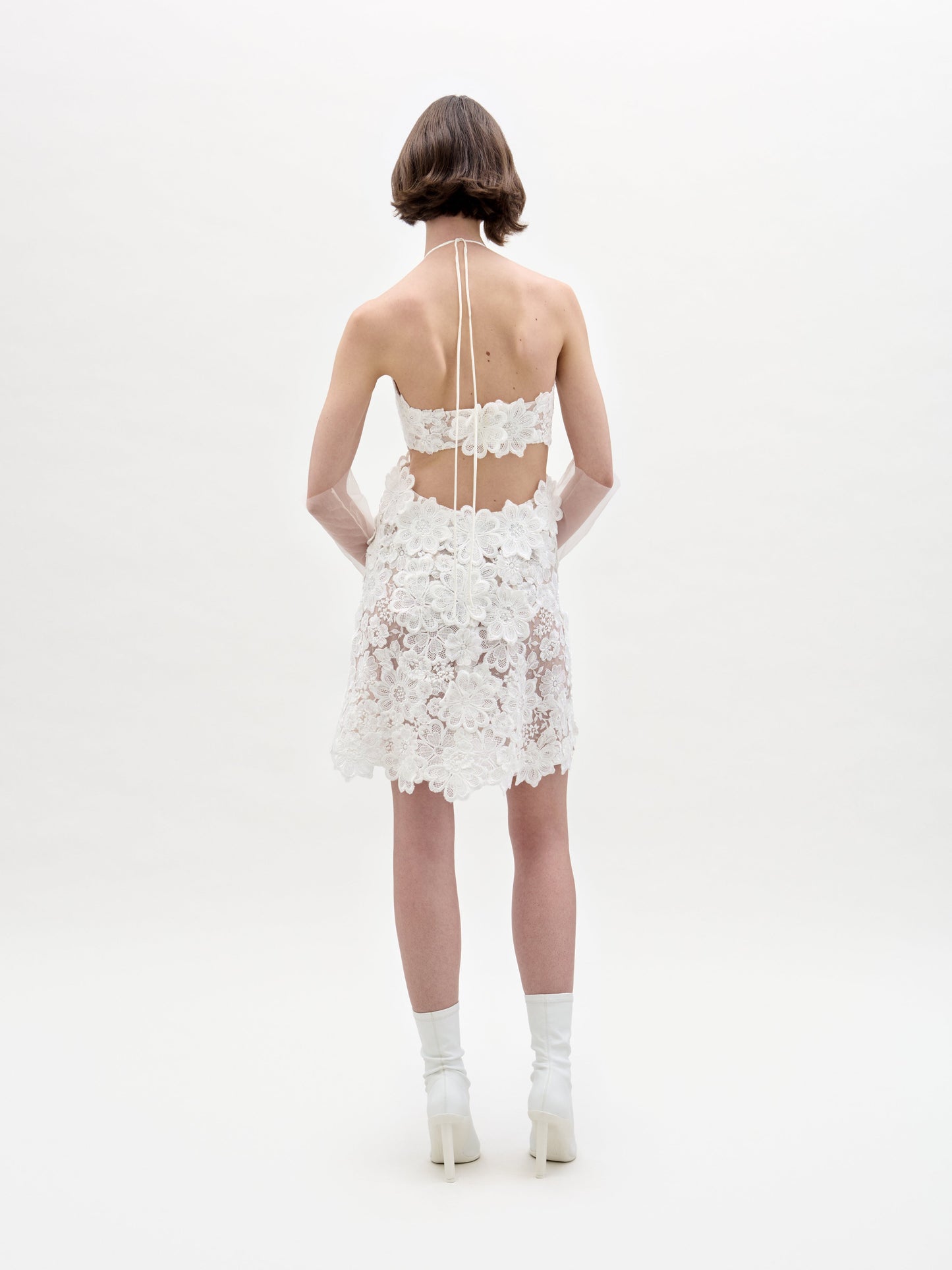 Una persona de cabello castaño corto está de espaldas a la cámara con un vestido Garazi, un diseño de encaje blanco sin mangas ni espalda descubierta con estampados florales. Completa el look con botas blancas de tacón y guantes transparentes sobre un fondo blanco liso.