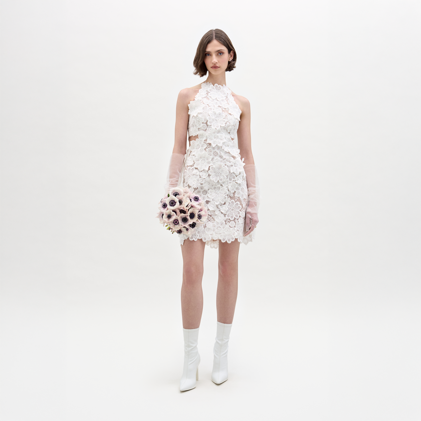 Una mujer aparece sobre un fondo blanco liso, vistiendo el Vestido Garazi, guantes transparentes, botines blancos y sosteniendo un ramo de flores de colores claros.