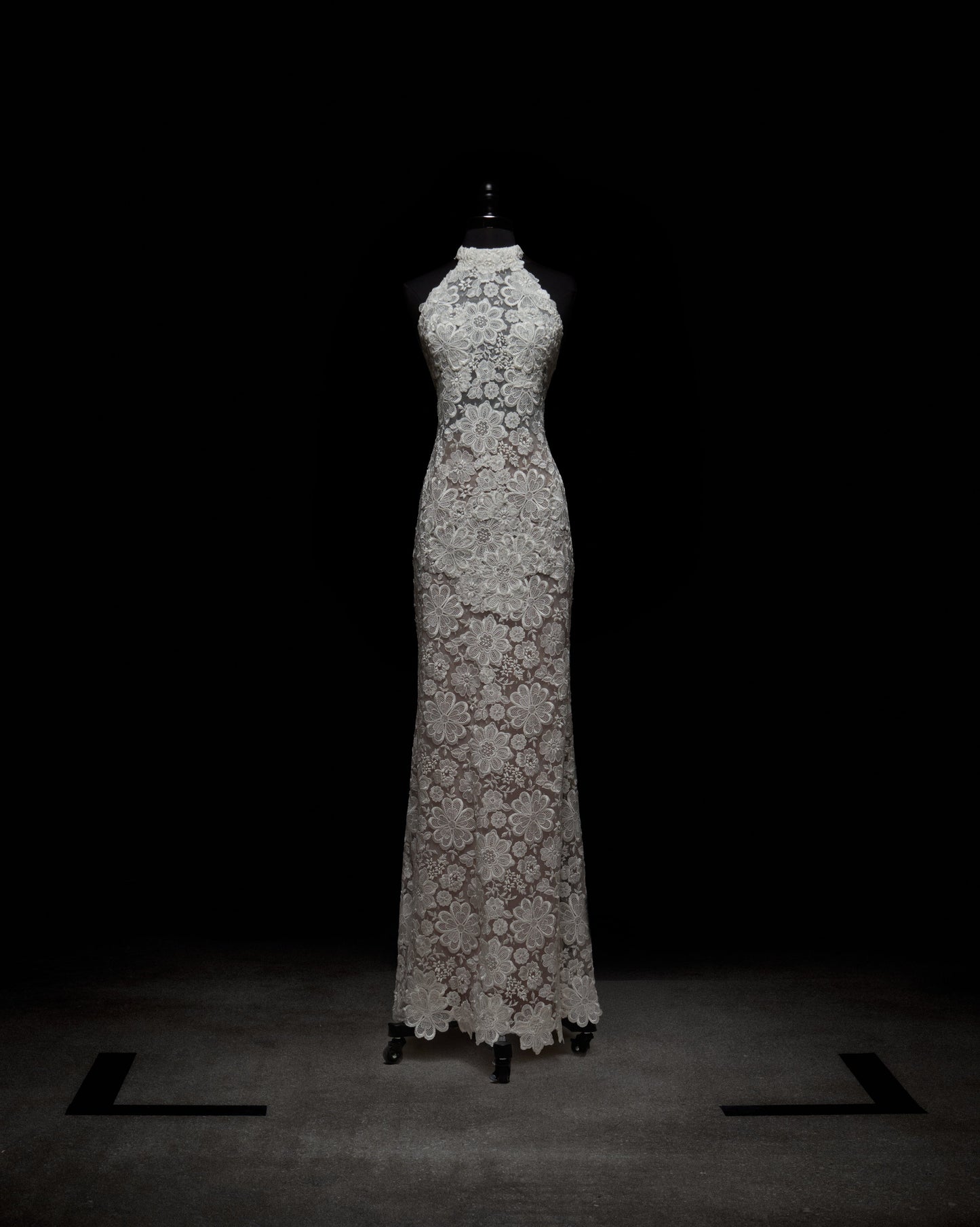 El Vestido Gardenia, un vestido de encaje blanco sin mangas hasta el suelo con escote alto, silueta ajustada y estampado floral, se exhibe en un maniquí negro sobre un fondo oscuro.