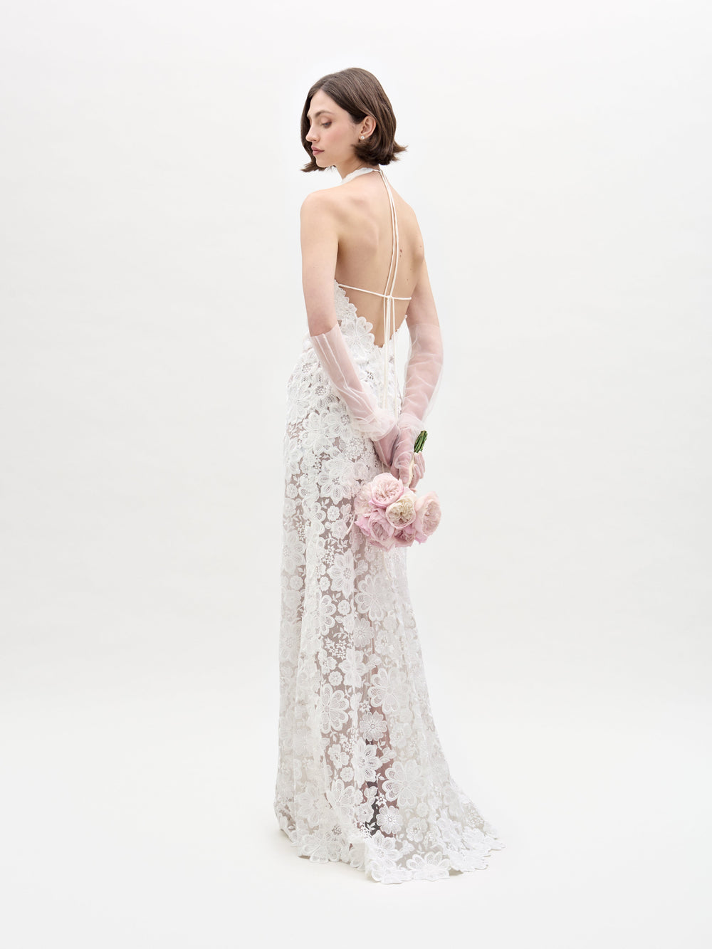 Una mujer que viste un Vestido Gardenia, un vestido de novia de encaje sin espalda y guantes transparentes está de pie mirando hacia otro lado, sosteniendo un ramo de rosas de color rosa claro detrás de su espalda contra un fondo blanco liso.