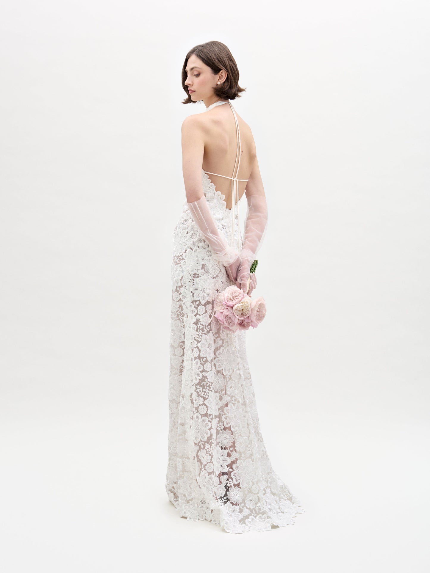 Una mujer que viste un Vestido Gardenia, un vestido de novia de encaje sin espalda y guantes transparentes está de pie mirando hacia otro lado, sosteniendo un ramo de rosas de color rosa claro detrás de su espalda contra un fondo blanco liso.