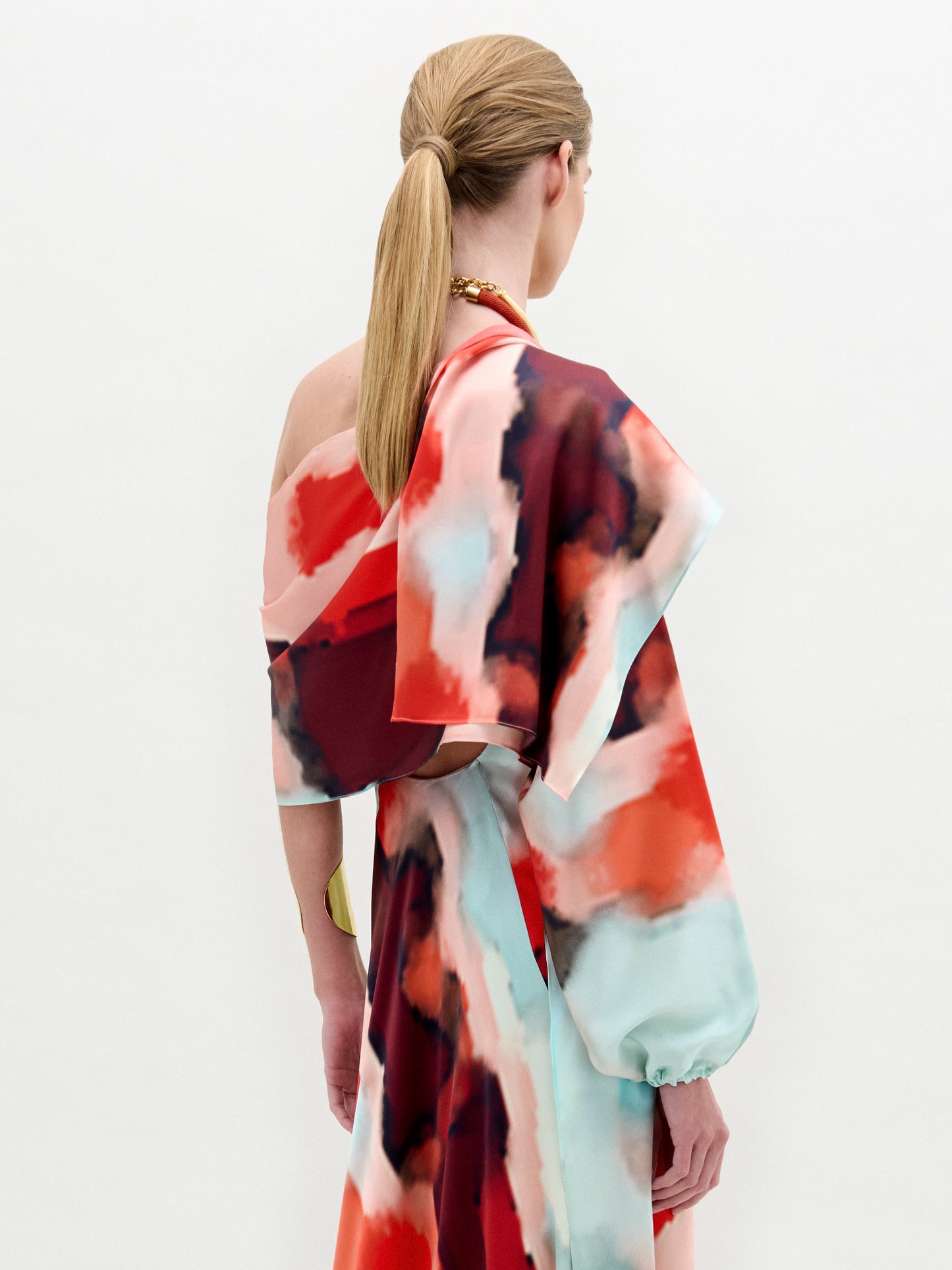 Una persona con el pelo liso, castaño claro y recogido en una coleta baja lleva el Gery Shrug -un colorido vestido con motivos abstractos en rojo, rosa, azul y blanco, confeccionado en tela italiana con manga asimétrica y espalda abierta- sobre un fondo liso.
