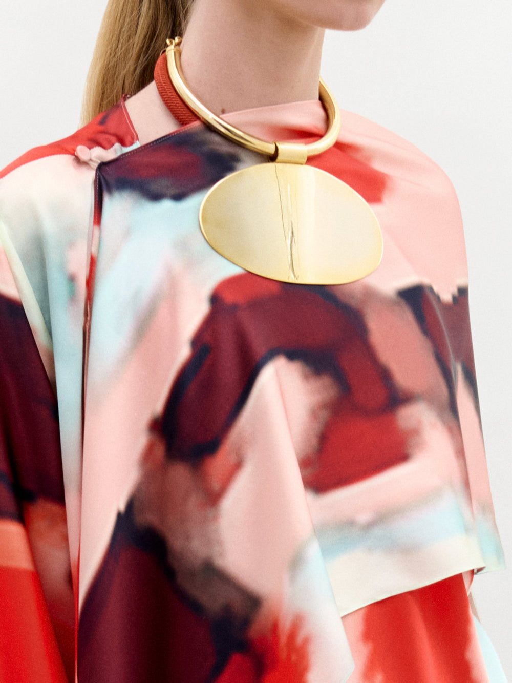 Sobre un fondo claro y liso, una persona luce el Gery Shrug, una prenda multicolor con motivos abstractos, combinada con un gran collar dorado con una banda gruesa y un colgante oval plano.