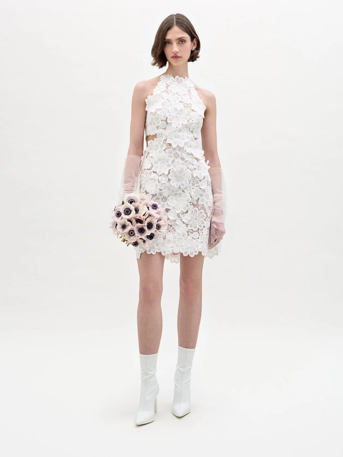 Una mujer se encuentra de pie sobre un fondo liso con el Vestido Garazi, un vestido corto de encaje blanco sin mangas con estampado floral. Lo combina con guantes transparentes, botines blancos y un ramo de flores rosa claro en la mano.