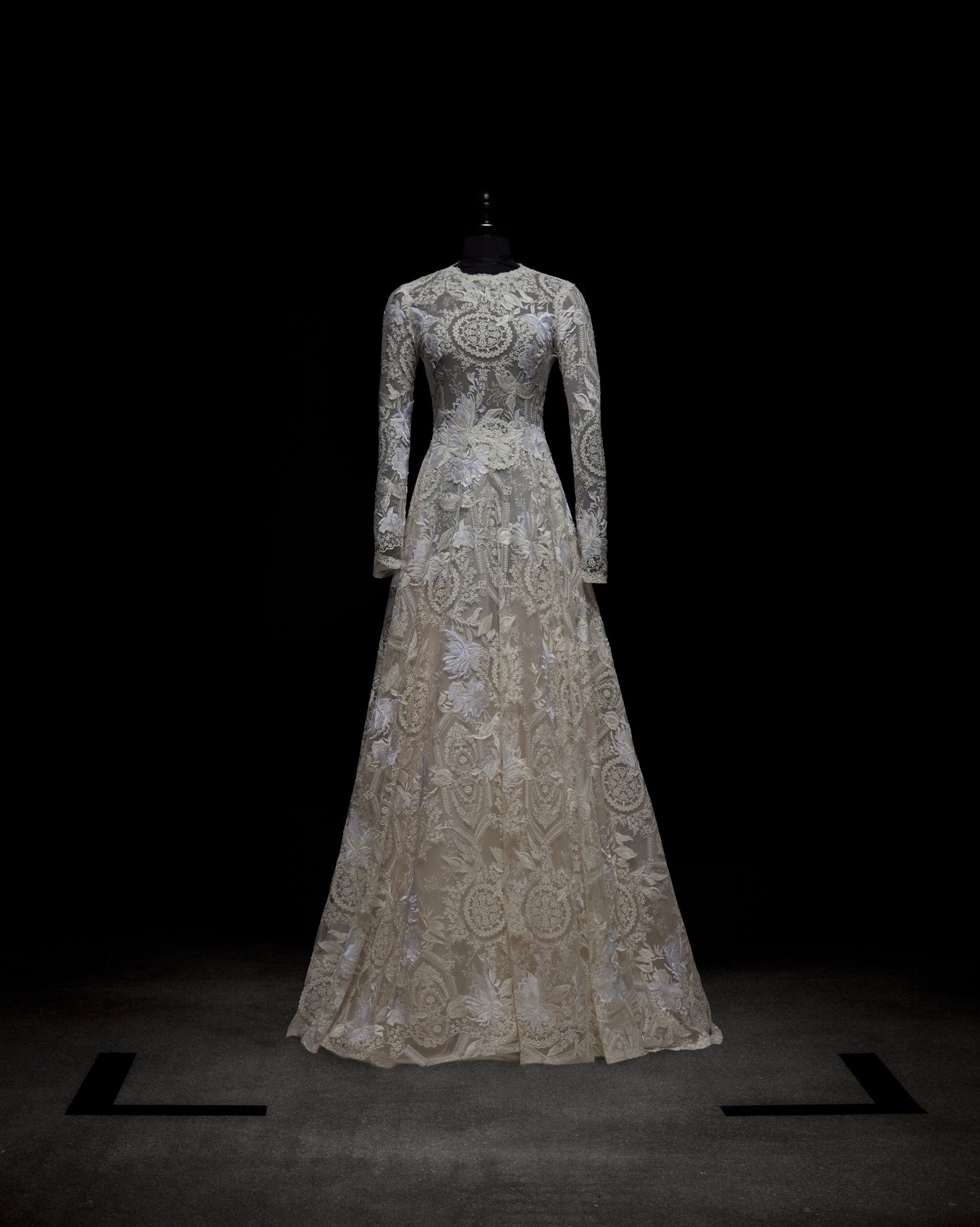 Exhibido en un maniquí negro sobre un fondo oscuro, el vestido Hermione es un vestido de encaje largo hasta el suelo, de manga larga, con patrones intrincados y un escote alto, que ofrece un aspecto elegante y clásico.