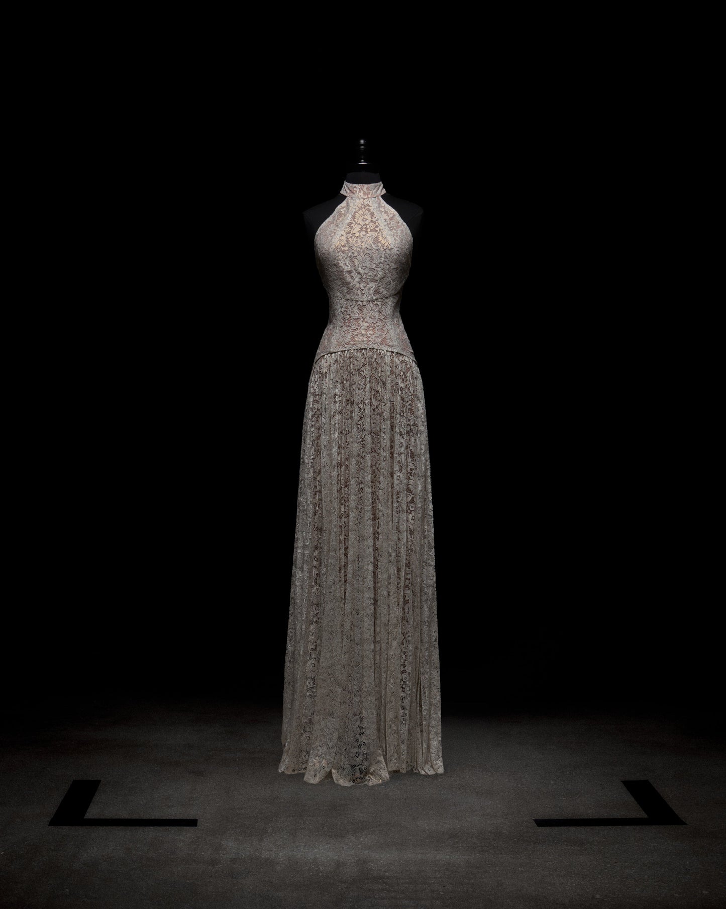 El vestido Ilana, un vestido de encaje largo hasta el suelo con detalles intrincados y un escote alto, se exhibe en un maniquí negro sobre un fondo oscuro, con una iluminación suave que resalta su diseño.