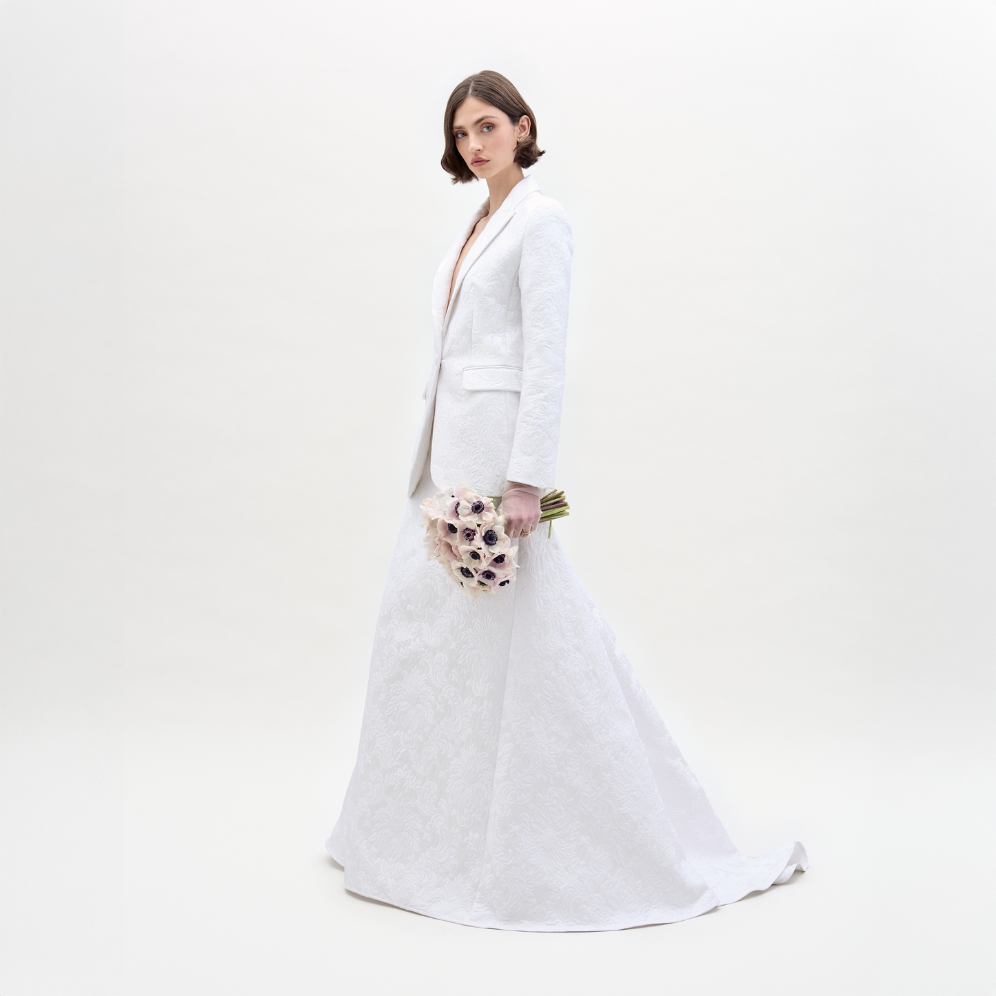 Con la Falda Imola, una falda larga blanca texturizada con silueta en forma de A y un blazer a juego, una persona está de pie sosteniendo un ramo de flores de colores claros sobre un fondo blanco liso.