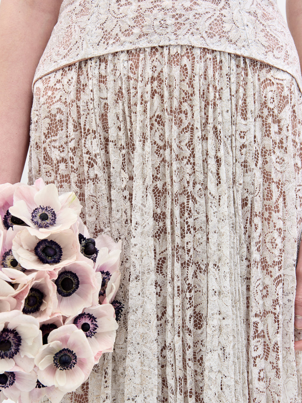 Una persona con un vestido Ilana, una creación de encaje en color hueso, sostiene un ramo de flores rosa pálido con centros oscuros. El estampado floral del vestido y la textura de las flores se fusionan para crear una apariencia delicada e intrincada.