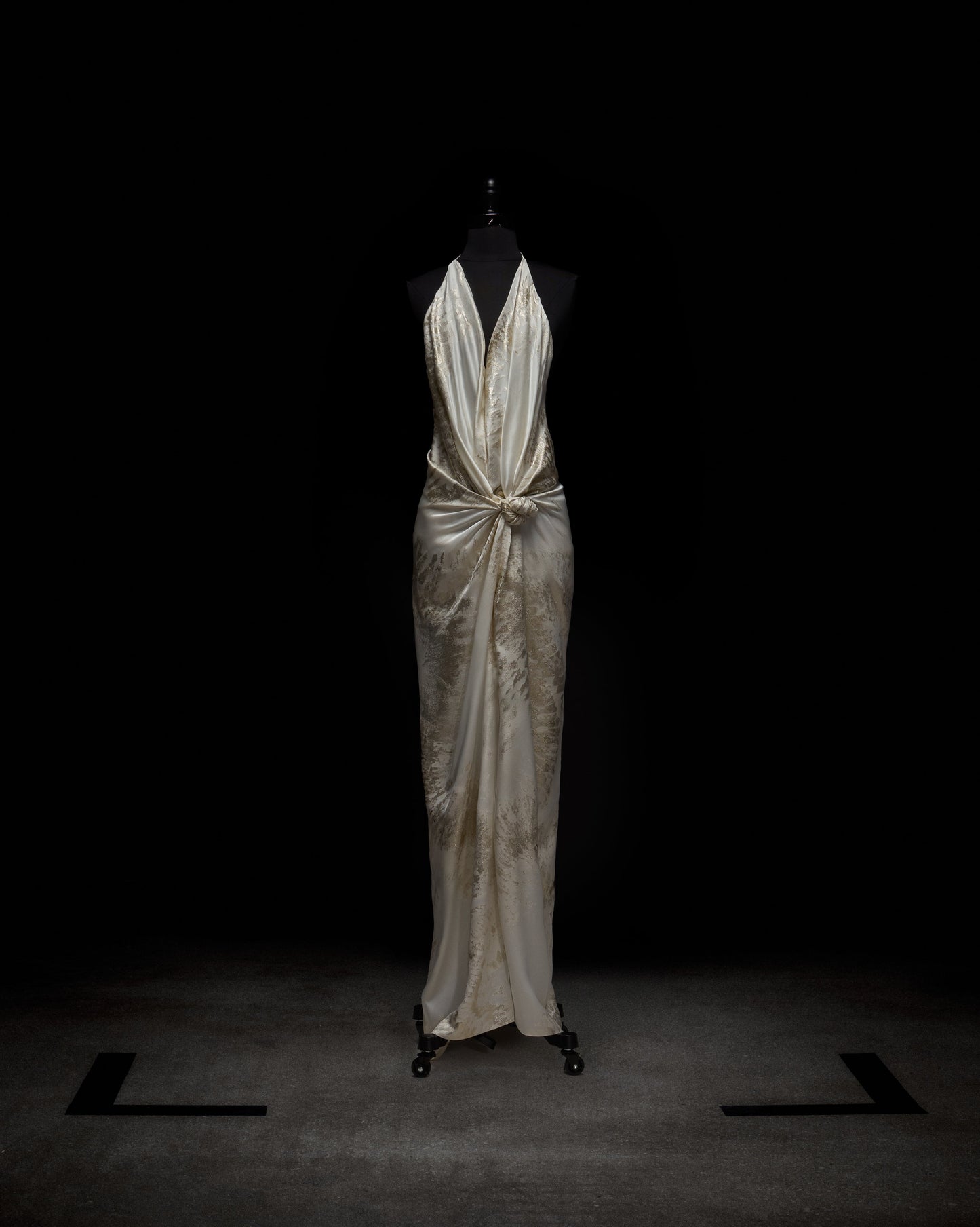 El vestido Jazmín se presenta en un maniquí sobre un fondo oscuro, con un acabado de satén blanco, cuello halter y silueta ajustada. Incluye un detalle fruncido en el centro delantero que se funde con una elegante falda larga.