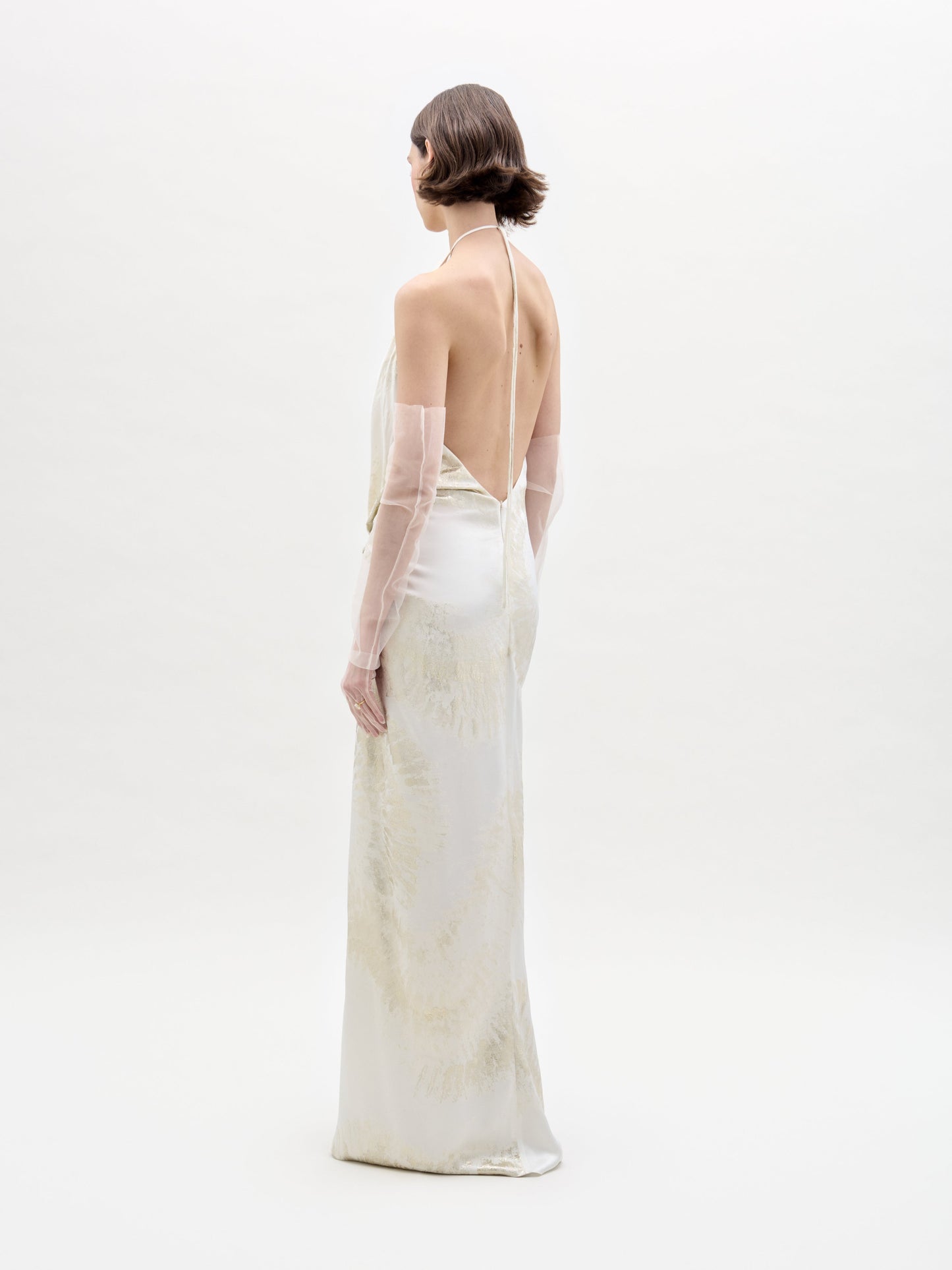 Una persona con cabello corto está de pie de lado sobre un fondo blanco, vistiendo el vestido Jazmin (un vestido blanco largo, sin espalda y con patrones sutiles e intrincados) combinado con guantes transparentes.