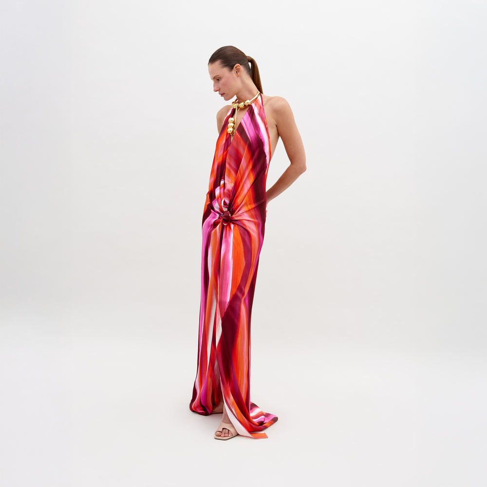 Una persona lleva el vestido Jazmin, un diseño largo y fluido con rayas verticales en rosa, naranja y morado, de pie sobre un fondo blanco. El vestido tiene un detalle de nudo en la cintura y la espalda abierta. El cabello peinado hacia atrás le suma estilo al conjunto.