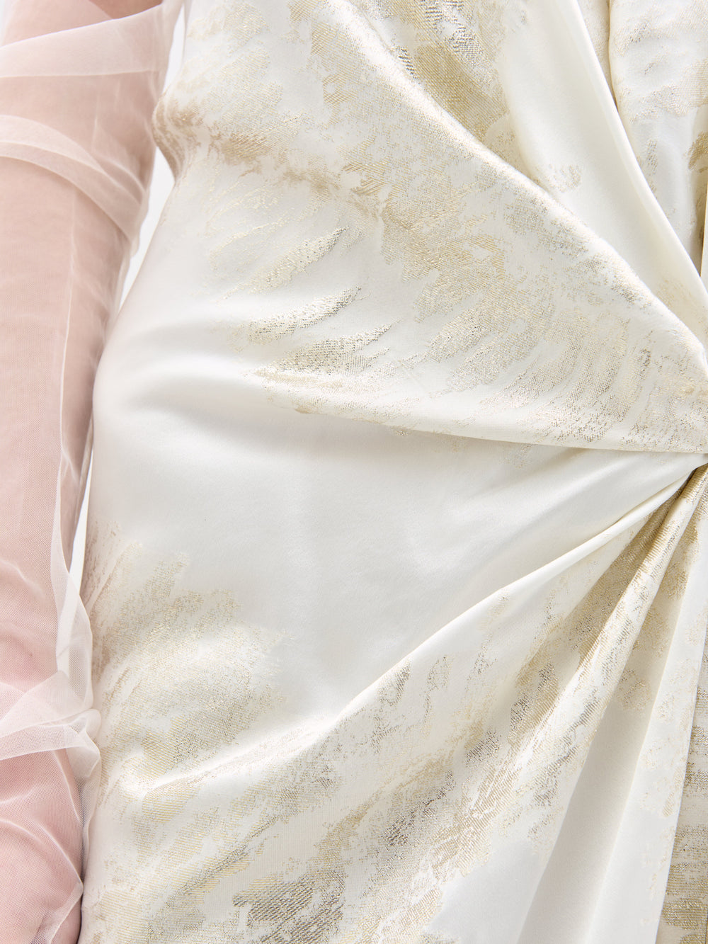 El vestido Jazmín presenta un patrón brillante y sedoso con textura blanca y dorada, con un diseño de nudo retorcido. Una manga transparente de color rosa claro deja al descubierto parcialmente un brazo en el lado izquierdo.