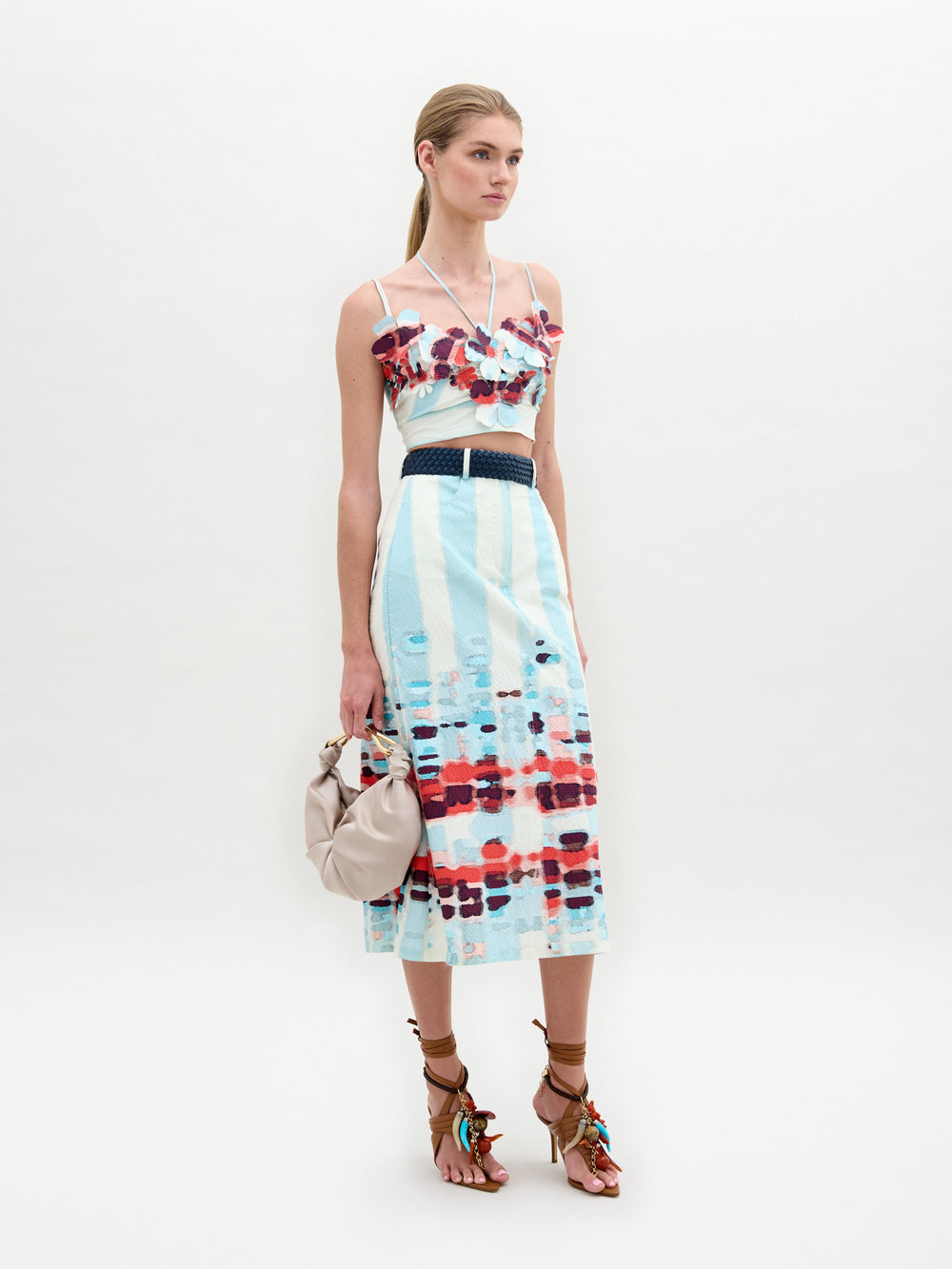 Una mujer está parada sobre un fondo liso con el vestido sin mangas Falda Layan, con estampados en azul, blanco y rojo confeccionado en tela italiana. Sostiene un bolso beige y usa sandalias marrones con borlas de colores. Lleva el cabello recogido.