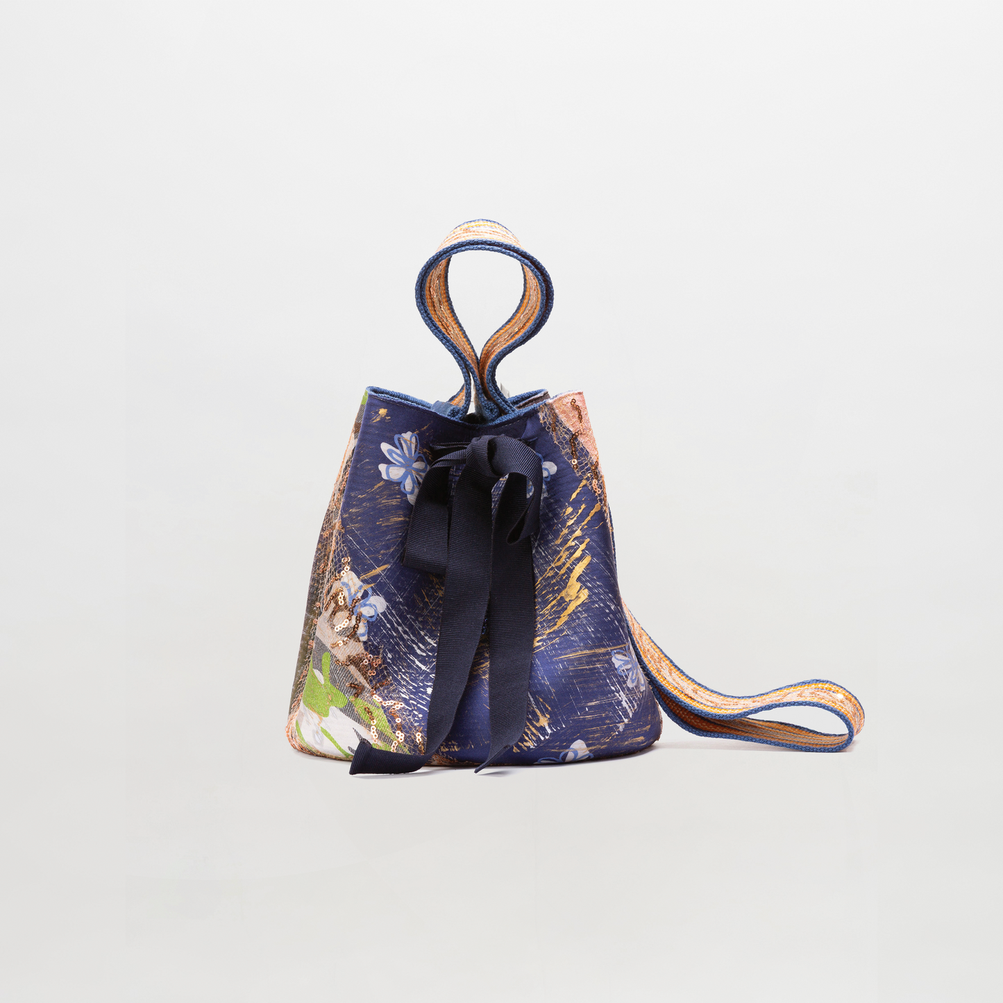 Mochila Laaput Big es un pequeño bolso tipo cubo con el exterior de tela azul, estampados de colores, un gran lazo azul oscuro en la parte delantera y una larga correa para el hombro, sobre un fondo blanco liso.