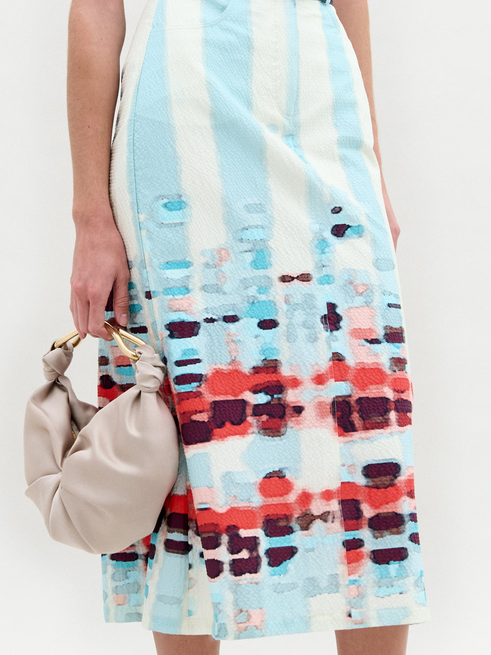 Una persona exhibe la Falda Layan, con un bolso de mano anudado de color beige claro. La falda midi, confeccionada en tela italiana, presenta un patrón gráfico con rayas verticales de color azul claro y detalles en rojo, granate, rosa, verde azulado y manchas azules.