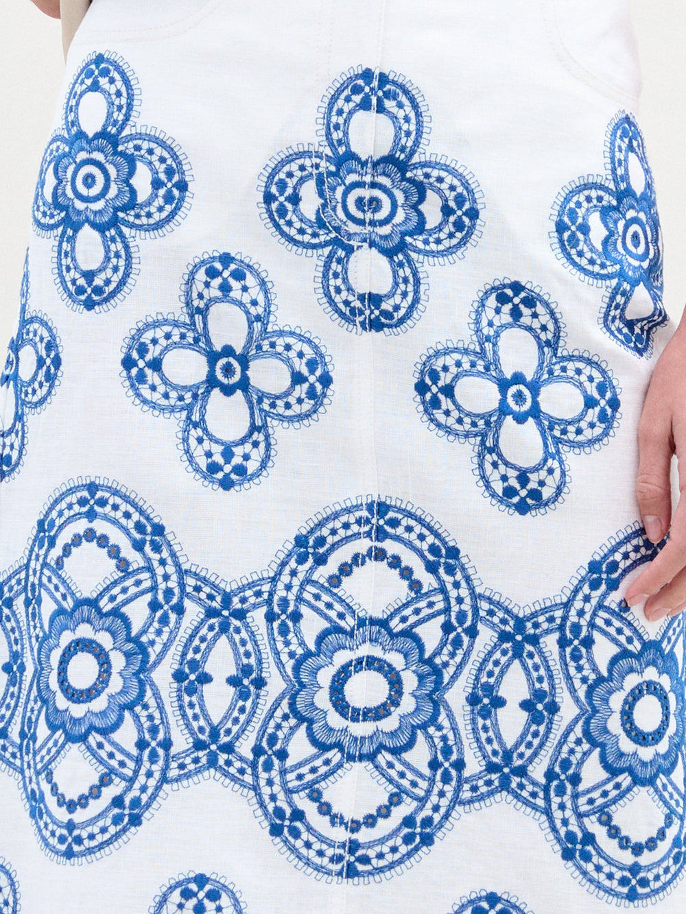 Primer plano de una persona que lleva la Falda Layan con bordados ornamentales azules, que recuerdan al lino bordado tradicional. Los intrincados patrones circulares y florales presentan formas simétricas que muestran una artesanía exquisita. Se puede ver parcialmente una mano en el lado derecho de esta pieza de Hecho en Colombia.