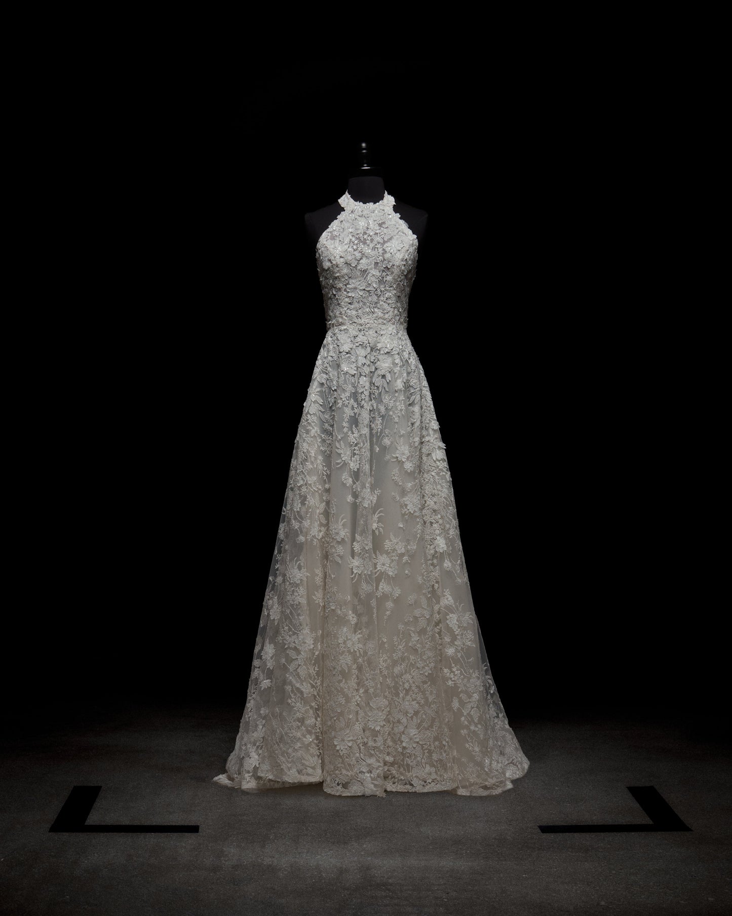 La escena, tenuemente iluminada, muestra el Vestido de Leia, un vestido de encaje blanco sobre un maniquí contra un fondo oscuro. El vestido resalta su corpiño ajustado, escote halter y falda larga adornada con intrincados detalles de encaje.