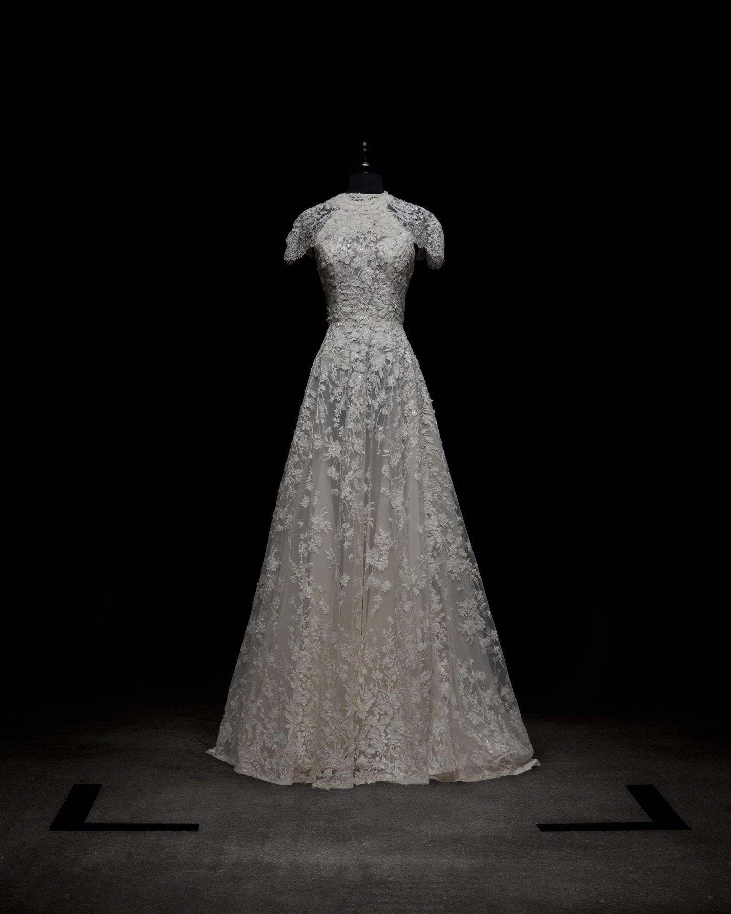 El Vestido Leia con Capa, un vestido de novia de encaje blanco hasta el suelo con mangas cortas, un corpiño ajustado y una falda acampanada, se exhibe en un maniquí negro sobre un fondo oscuro.