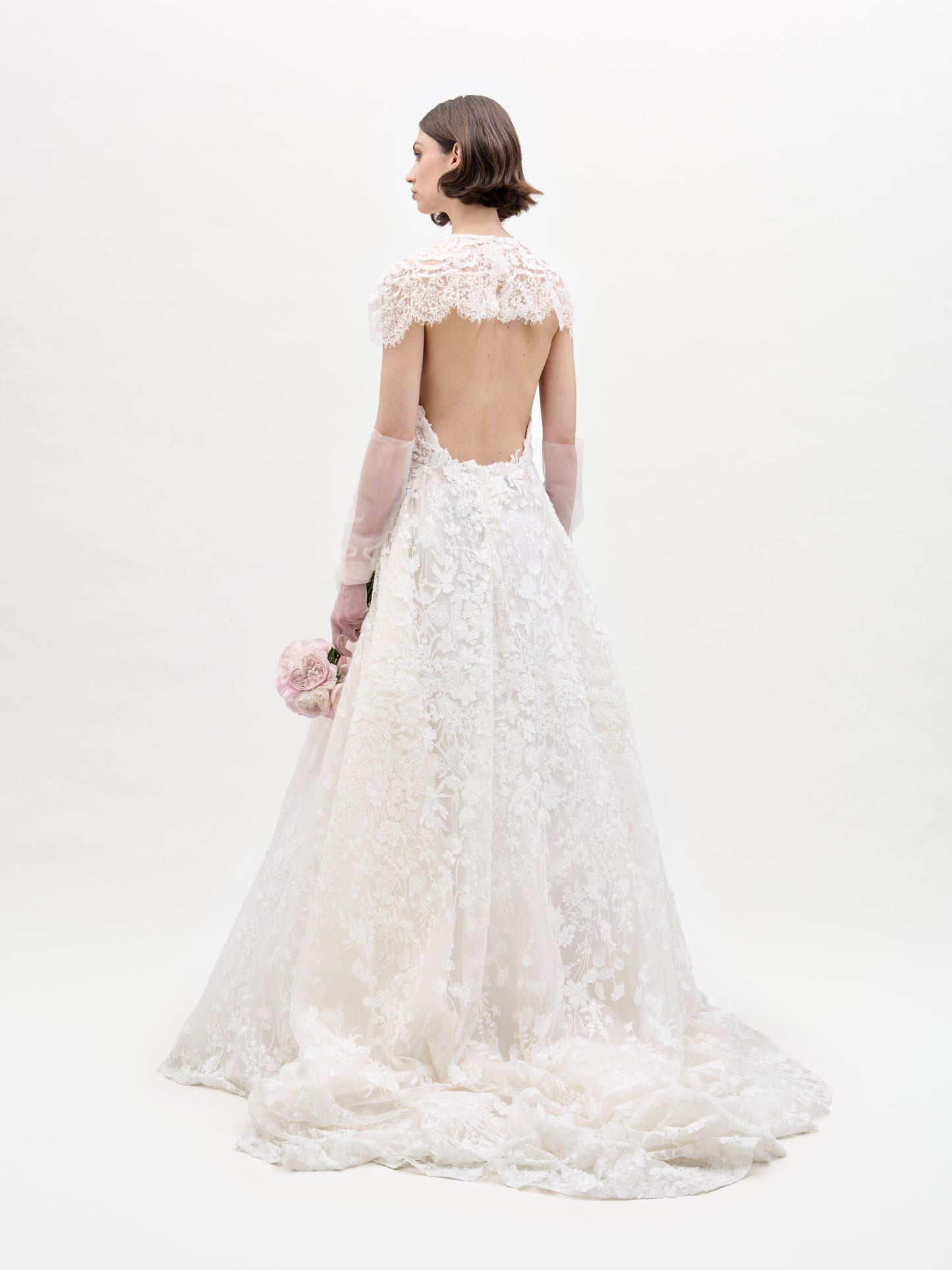 Una mujer mira hacia otro lado, vistiendo el Vestido Leia con Capa, un vestido de novia de encaje blanco largo hasta el suelo con espalda abierta y mangas largas transparentes, sosteniendo flores rosas y blancas sobre un fondo blanco liso.