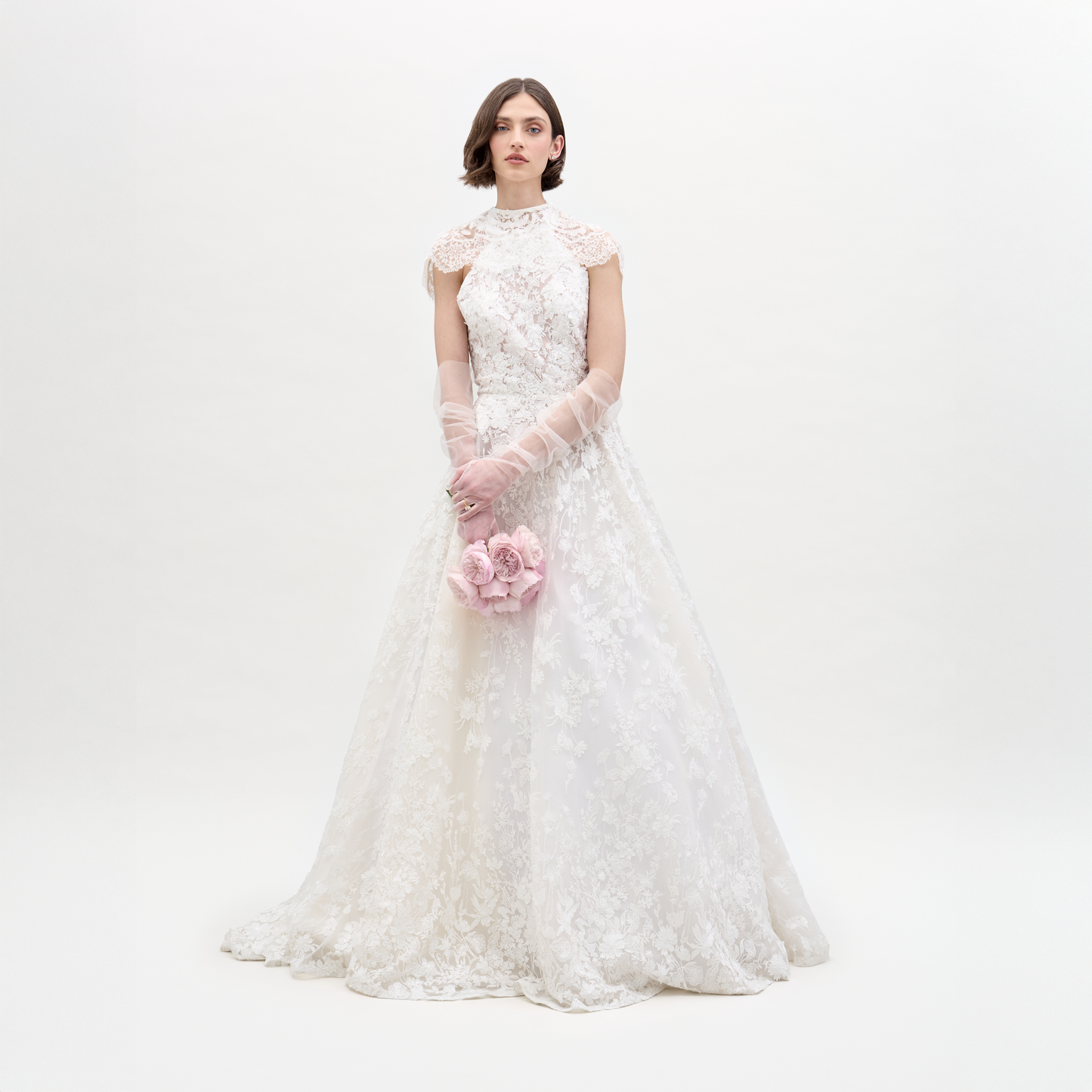 Una mujer con un vestido blanco de noche y guantes transparentes aparece de pie sobre un fondo liso y sostiene un pequeño ramo de flores de color rosa claro.