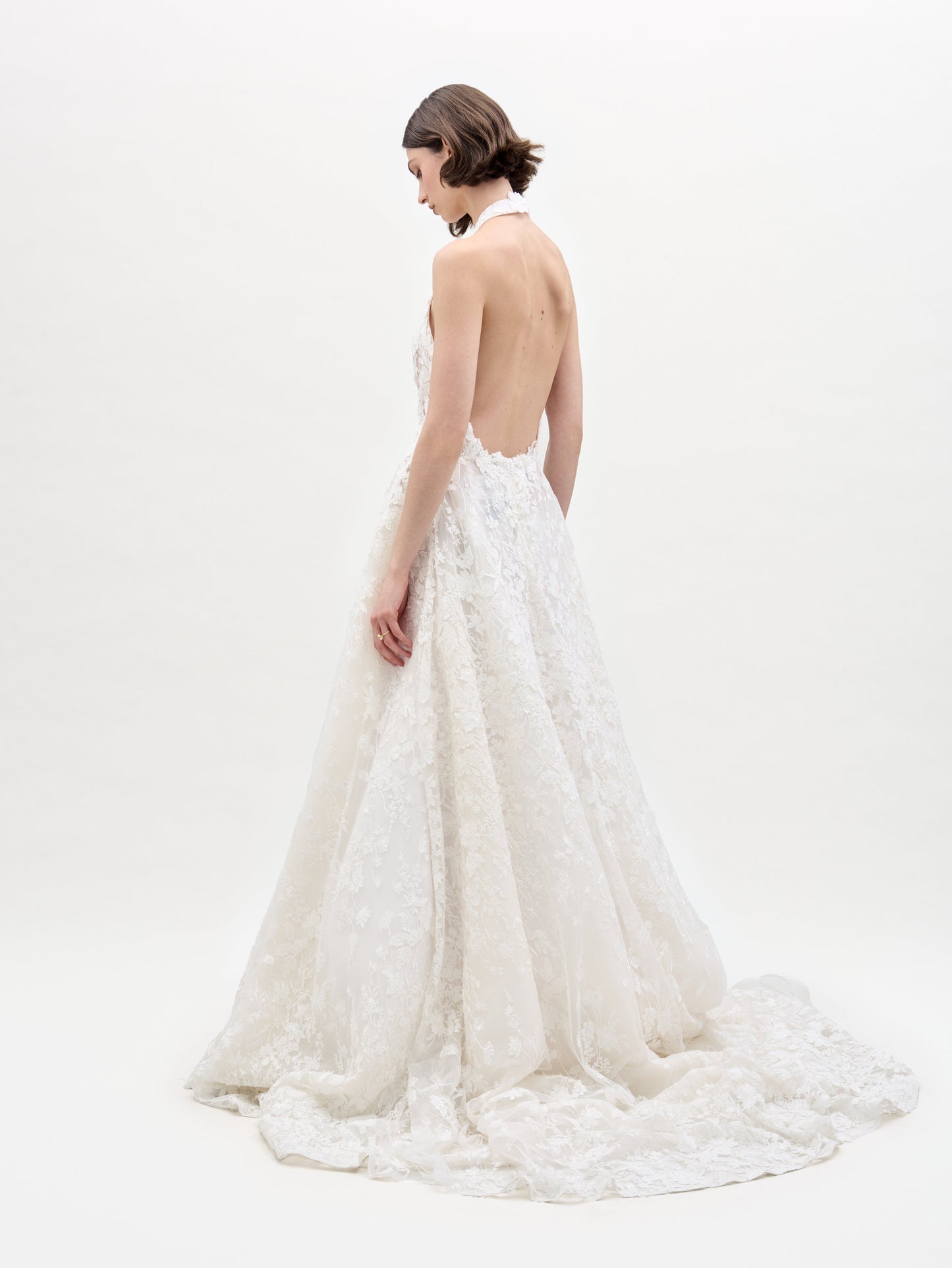 Una mujer de perfil luce el Vestido Leia, un vestido de novia de encaje blanco con cuello halter y espalda descubierta. Presenta una cola larga y fluida. Tiene el pelo corto y castaño y mira hacia otro lado, sobre un fondo blanco liso.