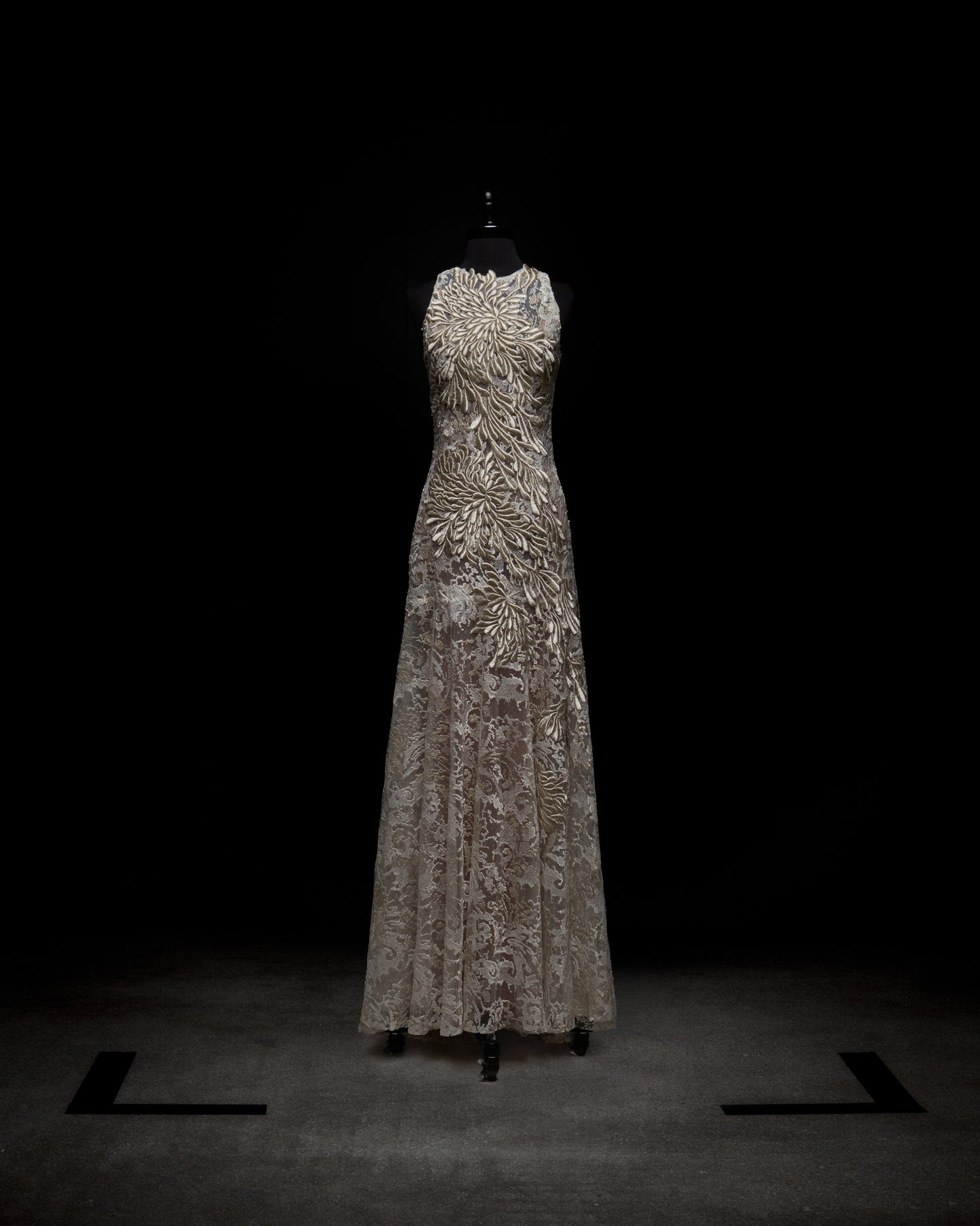 El vestido Levi, presentado en un maniquí negro sobre un fondo oscuro, luce un intrincado bordado en espiral dorado y plateado. Su corpiño ajustado se transforma en una falda ligeramente evasé hasta el suelo, ofreciendo una silueta elegante.