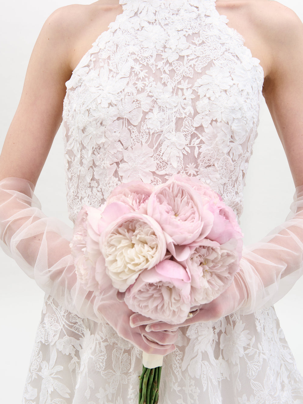 Una persona con el vestido de Leia, adornado con encaje floral, sostiene un ramo de peonías de color rosa claro y blanco mientras lleva guantes transparentes hasta el codo contra un fondo sencillo y de color claro.
