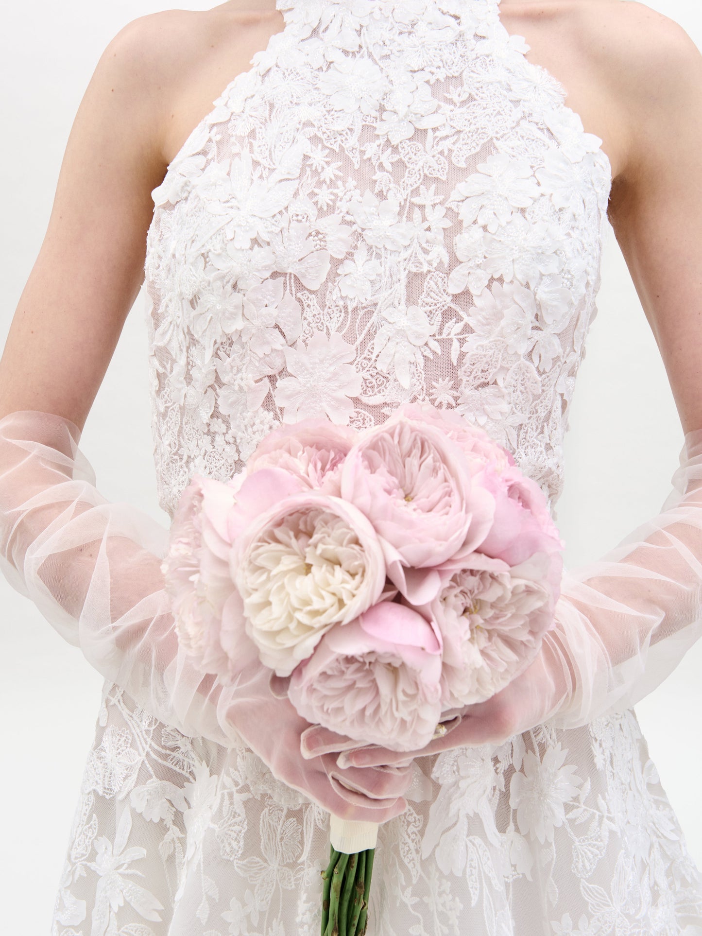 Una persona con el vestido de Leia, adornado con encaje floral, sostiene un ramo de peonías de color rosa claro y blanco mientras lleva guantes transparentes hasta el codo contra un fondo sencillo y de color claro.