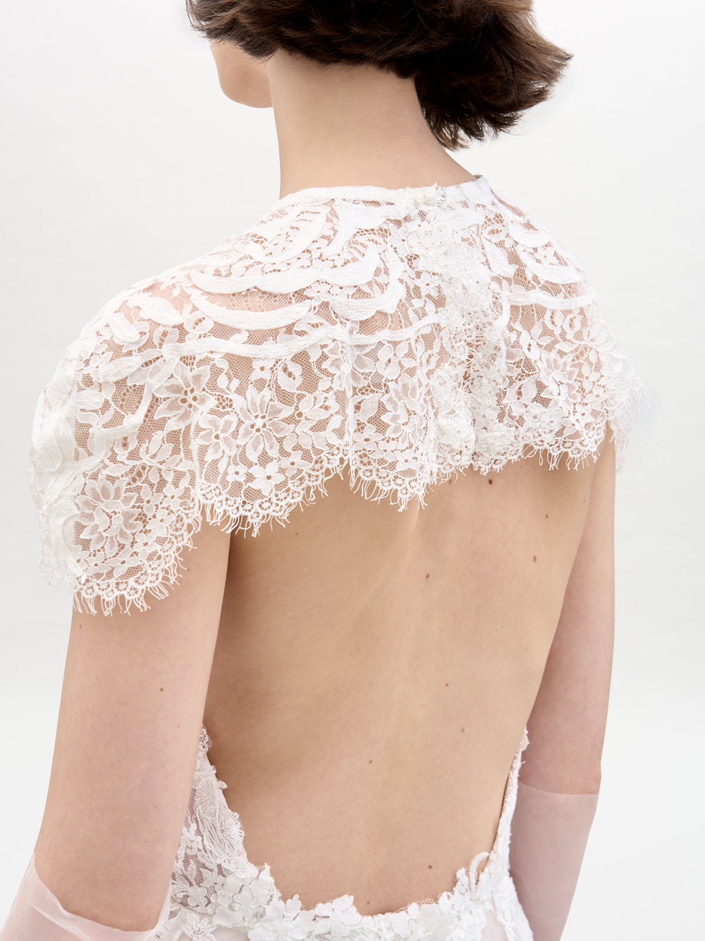 Se muestra a una persona con cabello castaño corto de espaldas vistiendo el Vestido Leia con Capa, un vestido de encaje blanco con espalda abierta y detalles intrincados de encaje floral en los hombros y la parte superior de la espalda, sobre un fondo liso de color claro.