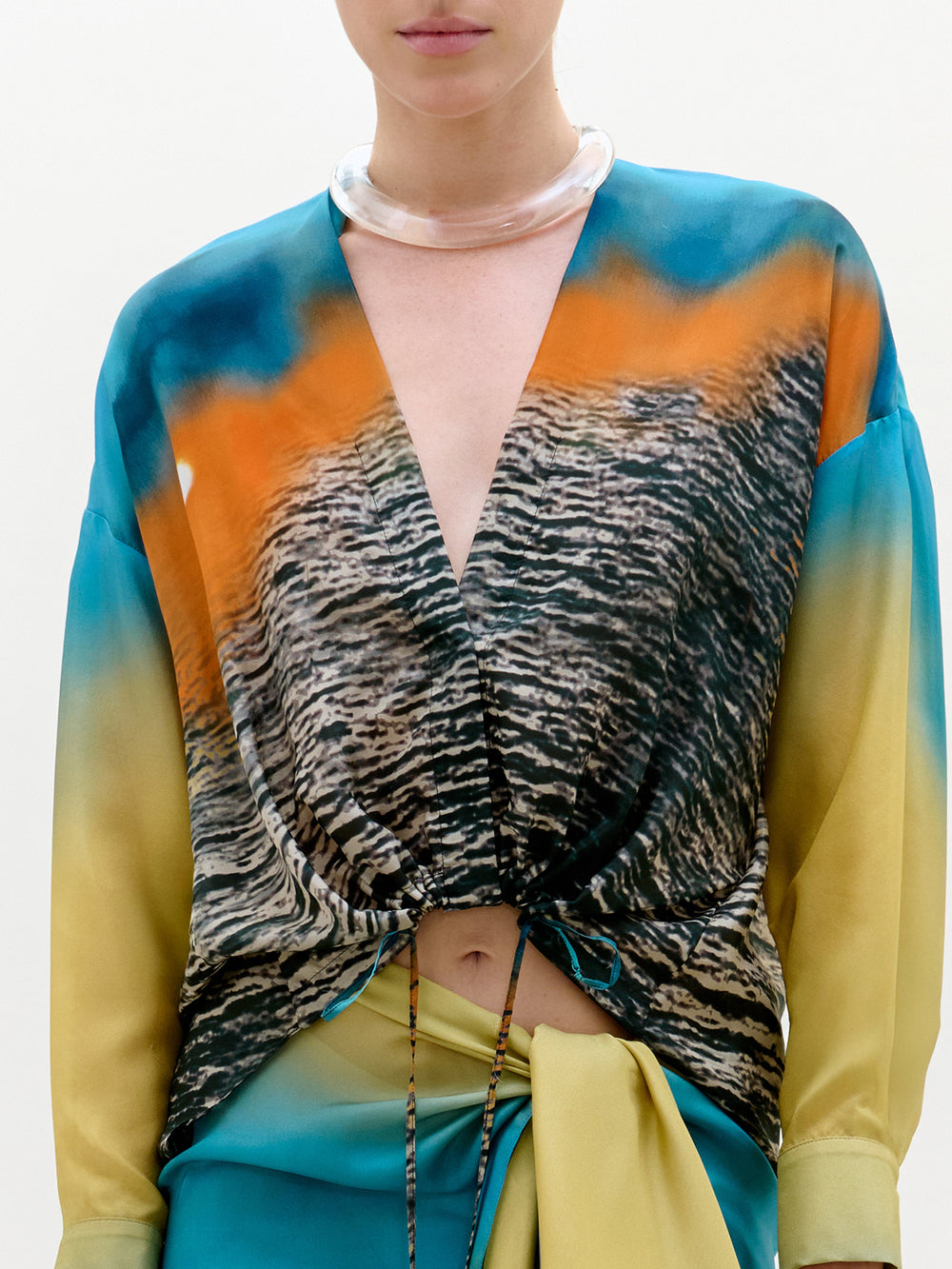 Una persona lleva la blusa Mérida -fabricada en Colombia, con un profundo escote en V, mangas largas y un estampado abstracto de agua y atardeceres en azul, naranja y amarillo- combinada con una gargantilla transparente. El rostro está parcialmente fuera del encuadre.