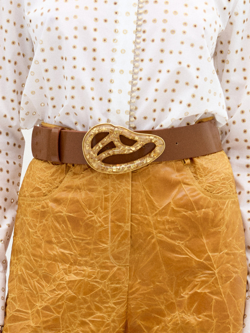Una persona lleva una blusa blanca de lunares metida por dentro de unos pantalones de cintura alta de color tostado con textura y el cinturón Nala, con una hebilla abstracta de Petralux.
