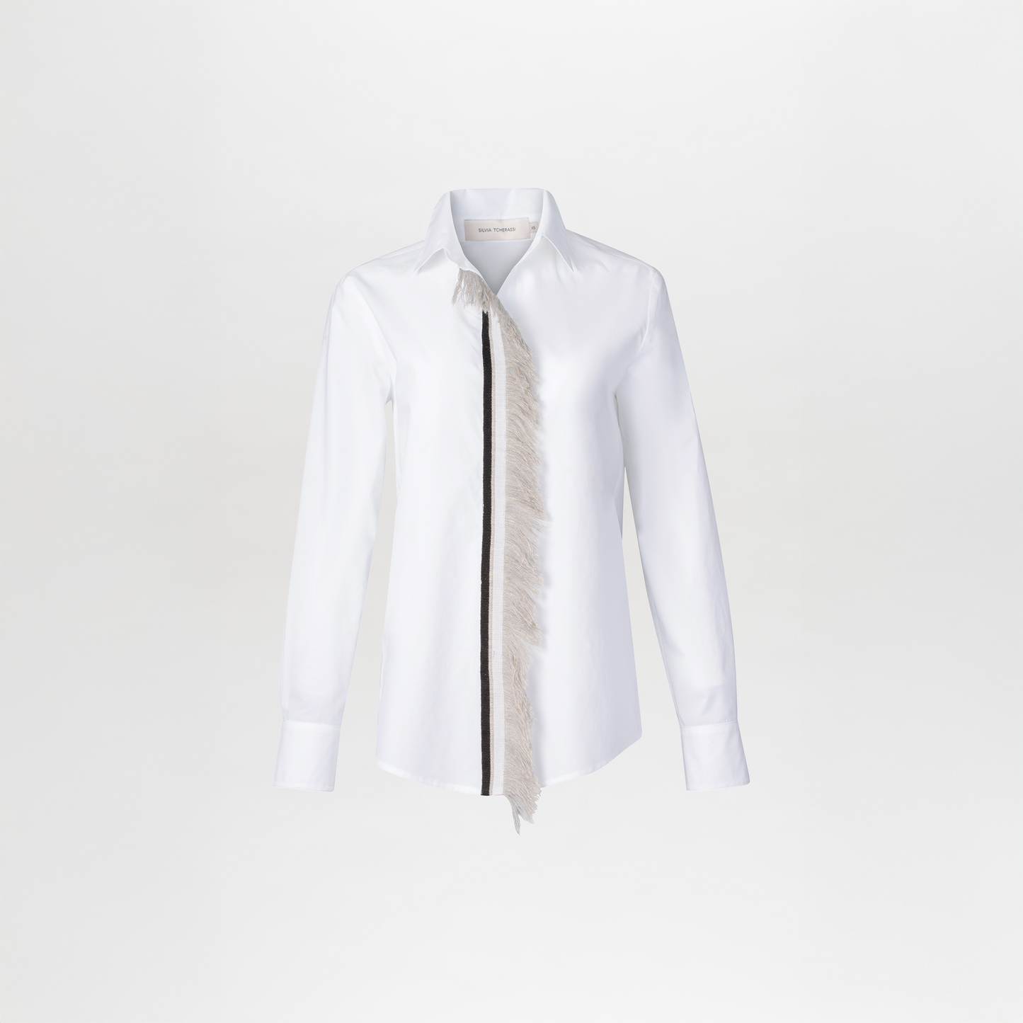 La Blusa Naroa es de algodón, manga larga y cuello clásico, con fleco en el borde y una franja negra asimétrica a lo largo de un lado de la tapeta de botones.